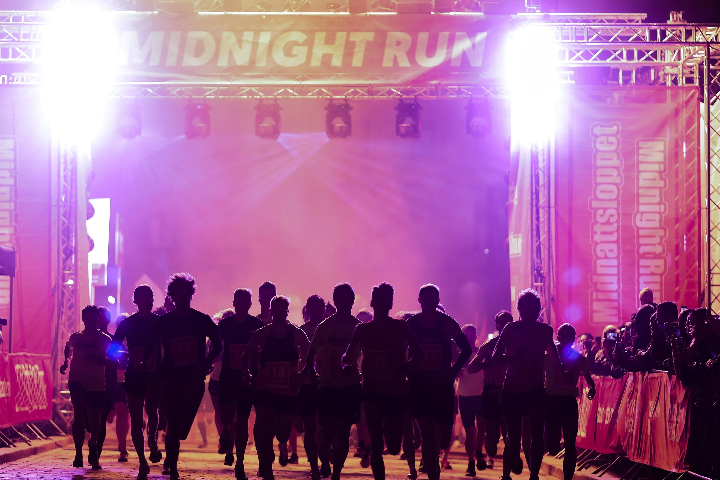 Runners at the start of a midnight marathon race at night, silhouetted under bright purple and pink lighting, with a large banner above reading 'MIDNIGHT RUN' and event branding along the sides.