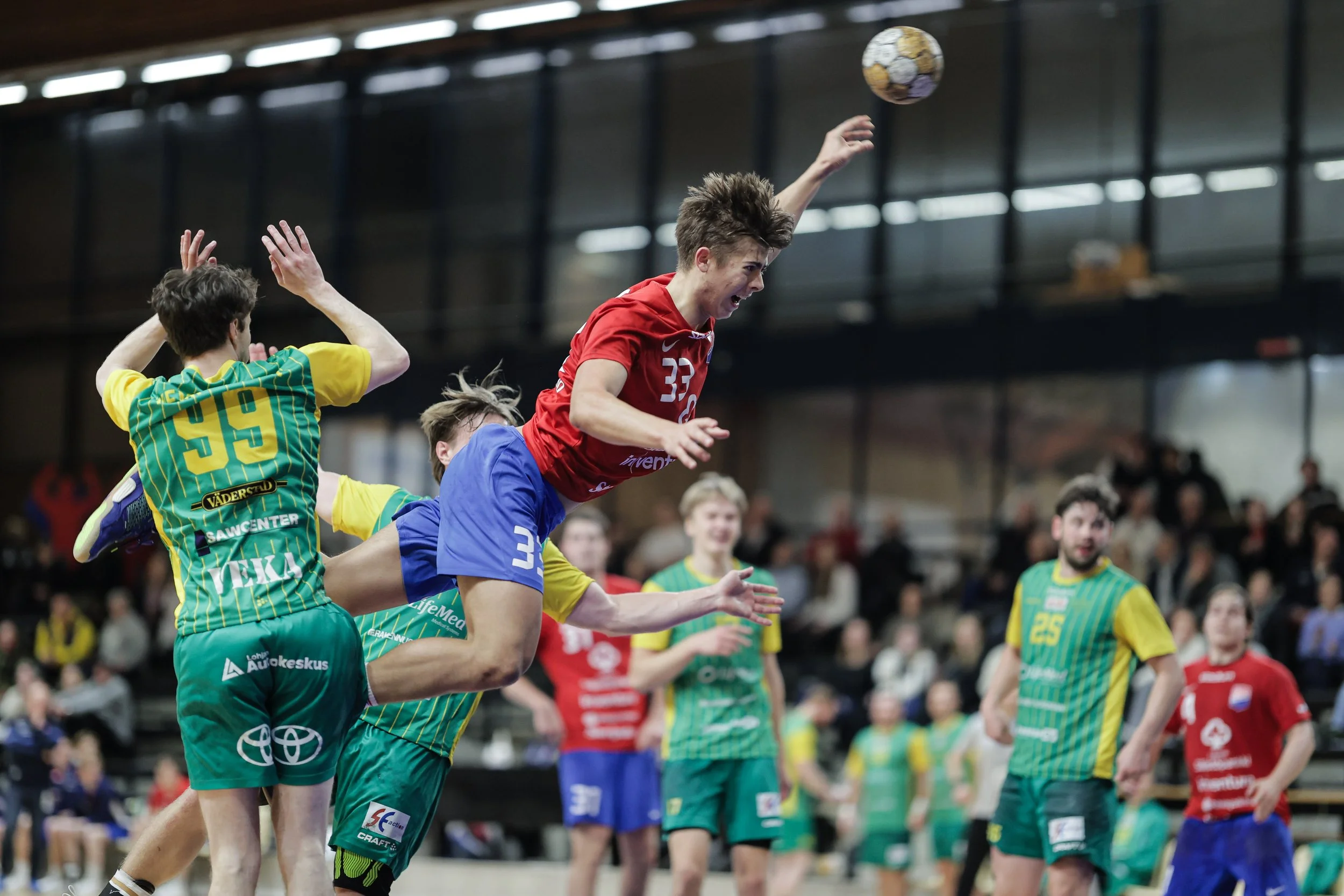 Young male handball player in red jersey jumping to shoot the ball, surrounded by players in yellow and green jerseys during a match in an indoor gymnasium.