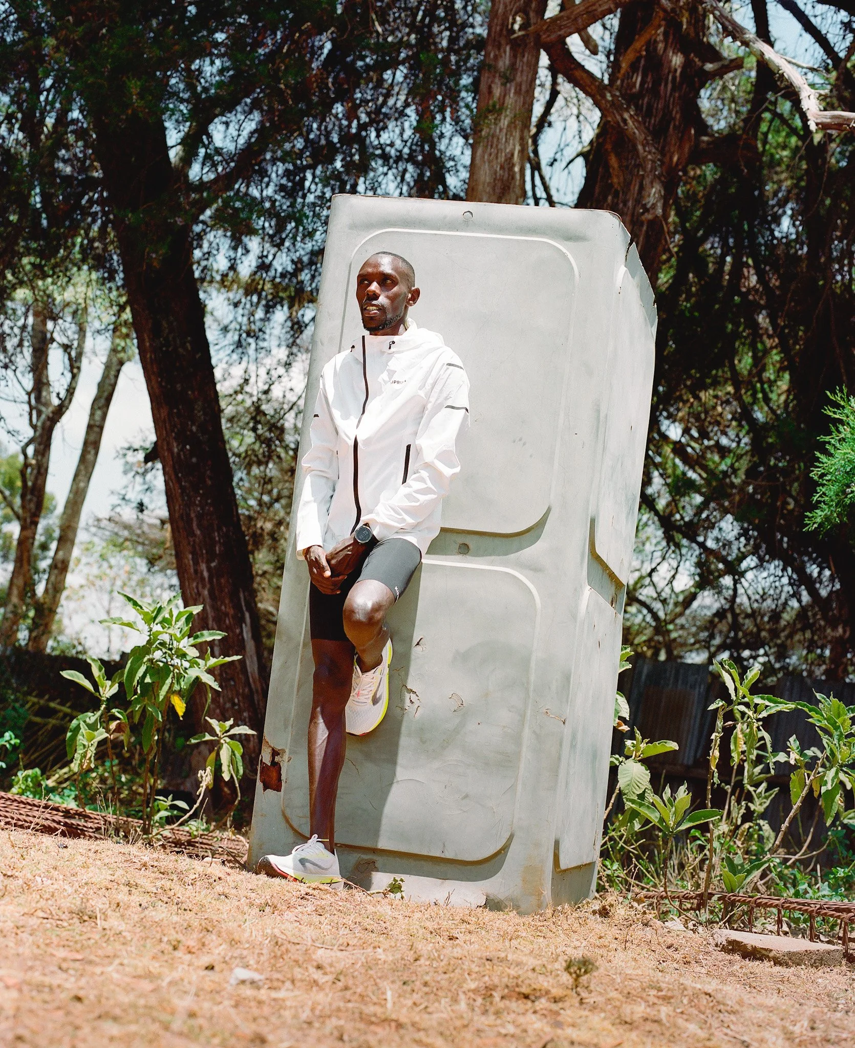 Un homme au sport, portant une veste blanche, des shorts noirs et des chaussures de course, suspend son pied sur une vieille cabine téléphonique en extérieur, avec des arbres en arrière-plan.