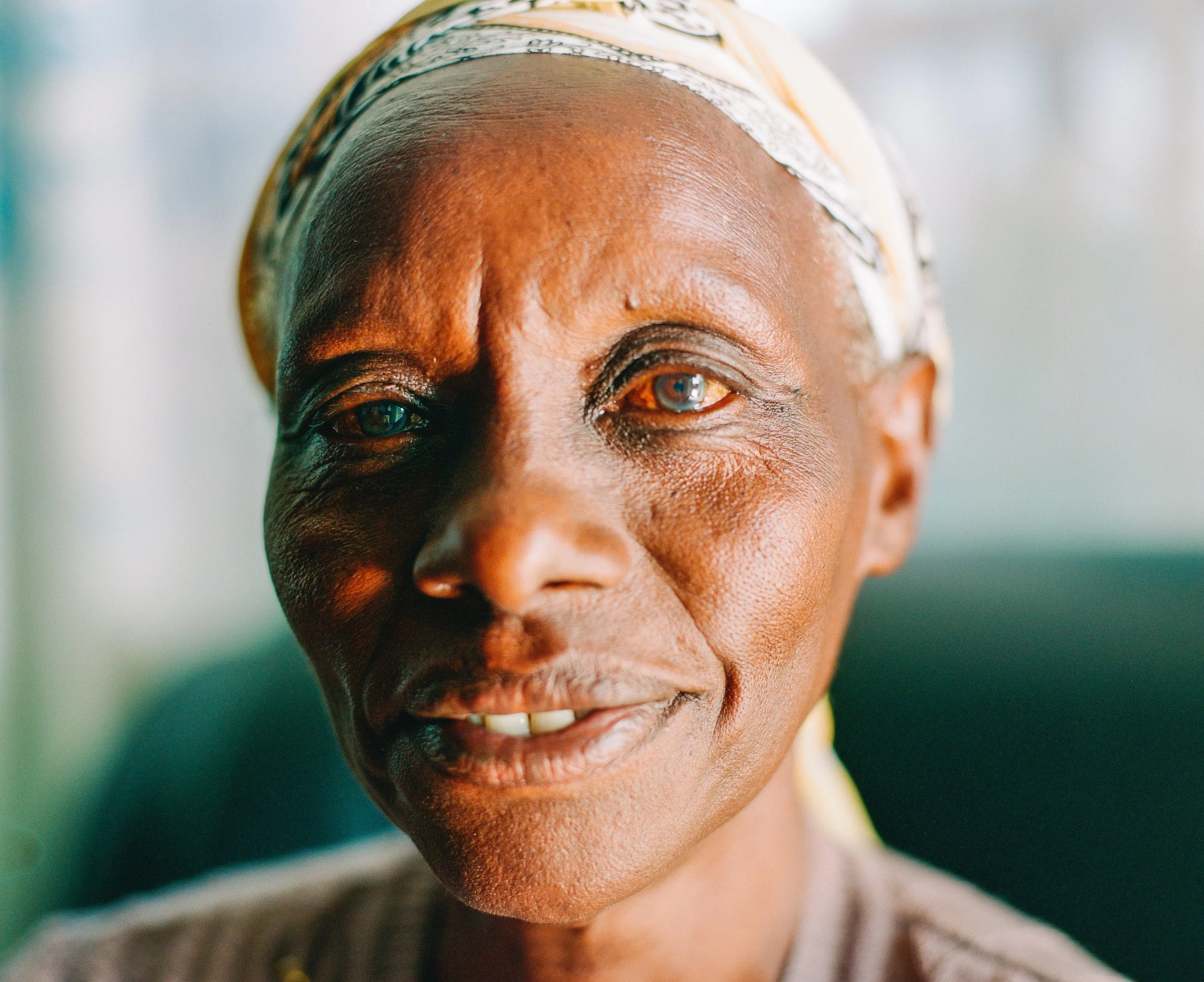 Portrait d'une femme âgée avec une peau foncée, portant un foulard blanc et beige, souriante et regardant vers l'objectif.