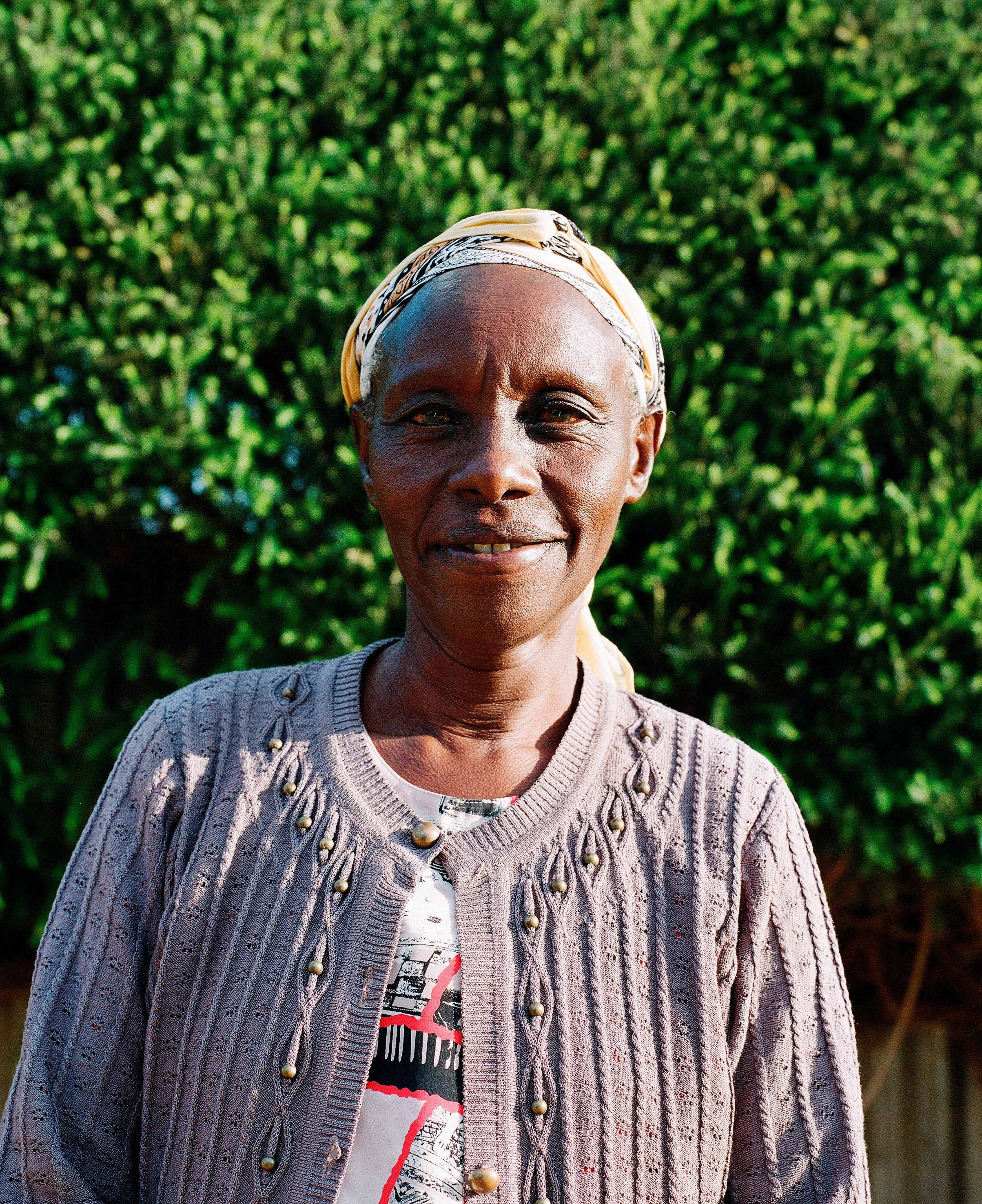 Une femme âgée porte un foulard beige noué autour de la tête, un pull beige orné de perles dorées et un t-shirt blanc avec un motif rouge, noir et blanc. Elle se tient devant un arrière-plan de verdure luxuriante, en plein soleil.