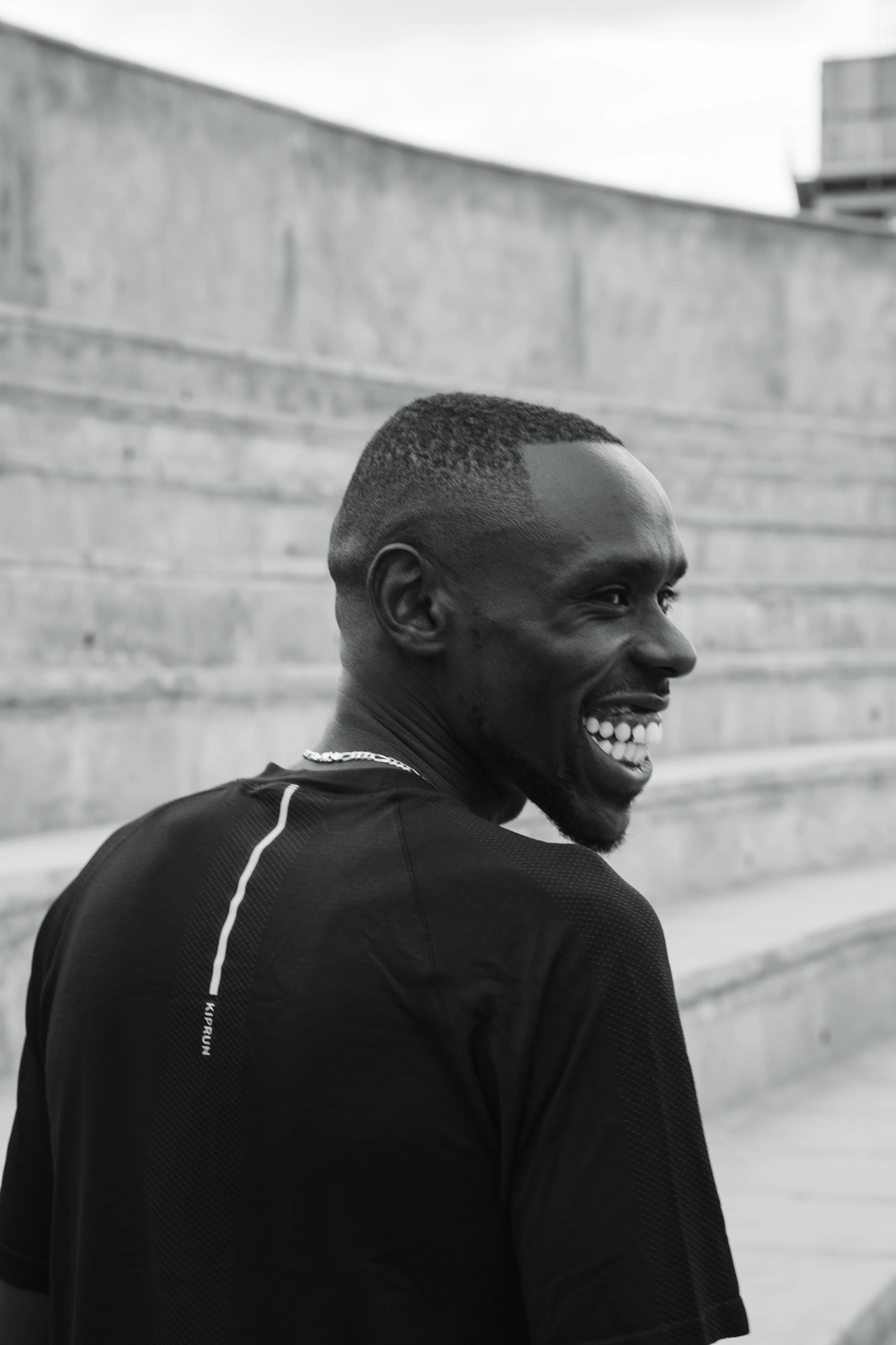 Homme souriant portant un maillot noir, en extérieur, avec des marches en béton en arrière-plan.
