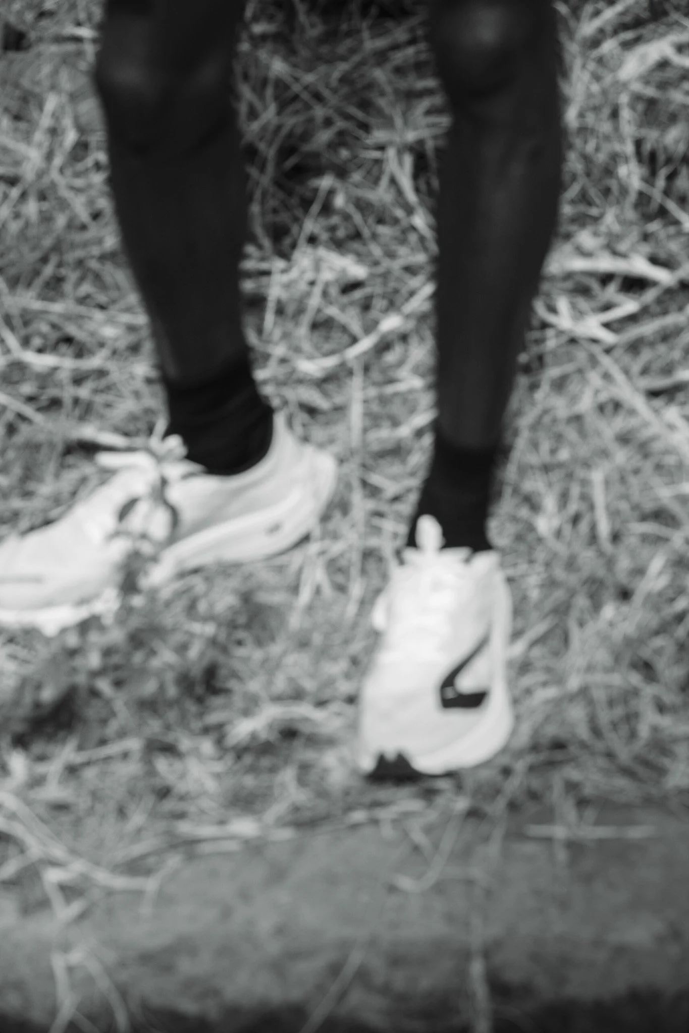 Vue floue des jambes d'une personne portant des chaussures de course blanches et un pantalon noir, avec un sol recouvert de végétation morte.