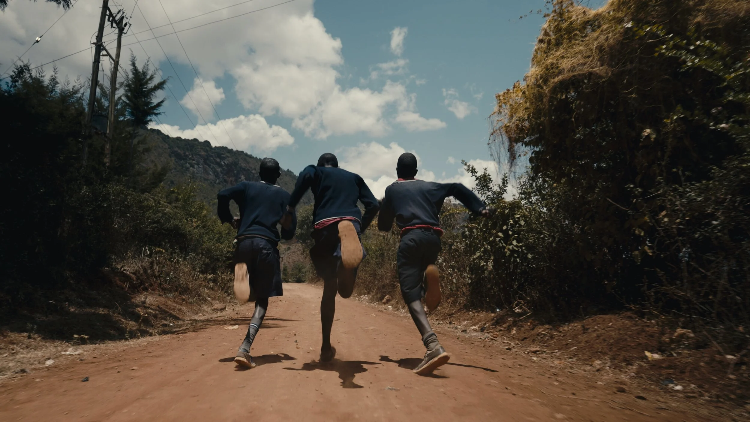 Trois jeunes coureurs en uniformes scolaires courant sur un chemin de terre en milieu rural, avec des montagnes, du ciel bleu et des nuages en arrière-plan.