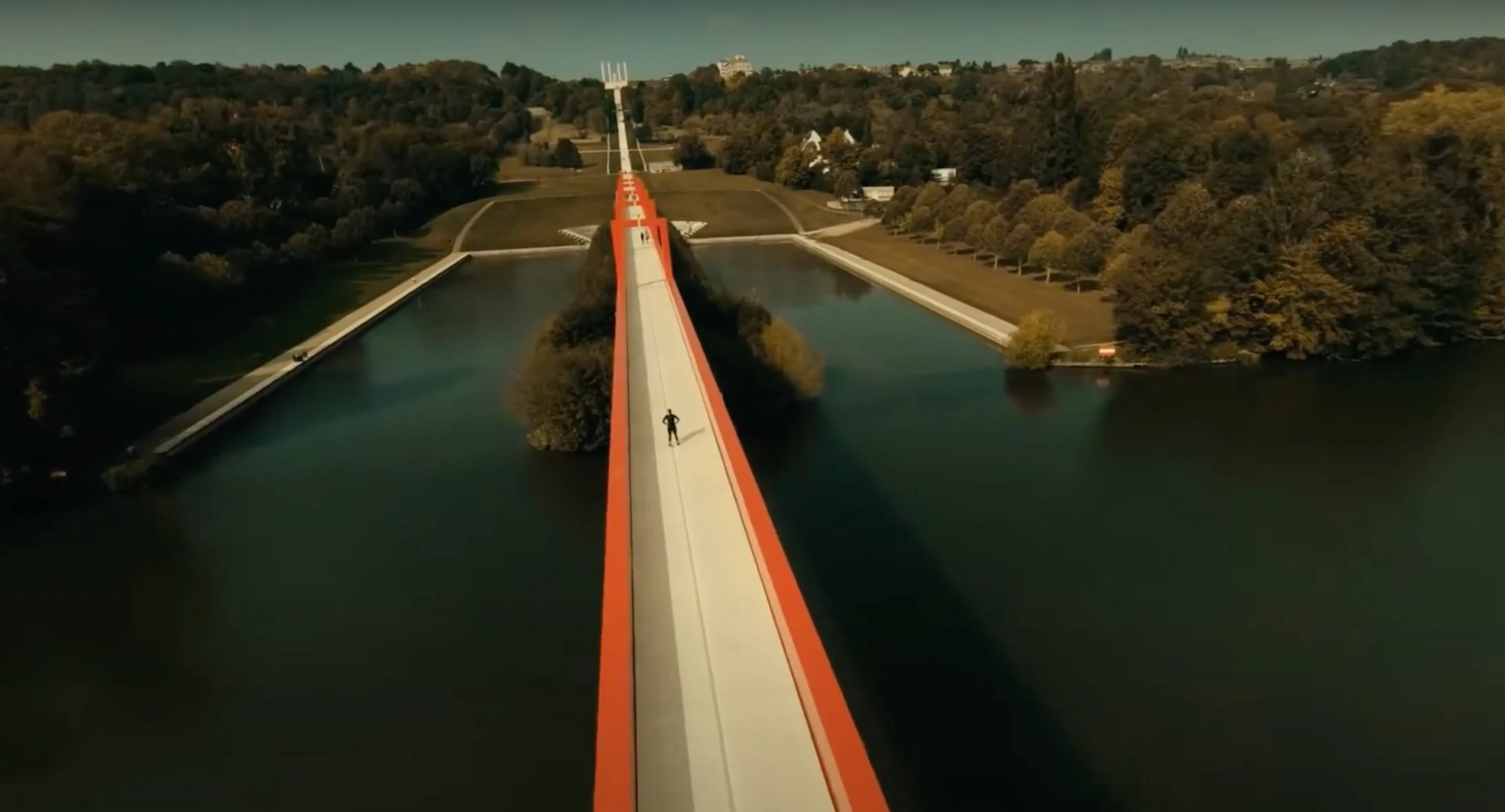 Un homme court sur une piste d'athlétisme suspendue au-dessus de l'eau, entourée d'arbres et de terrains verts.