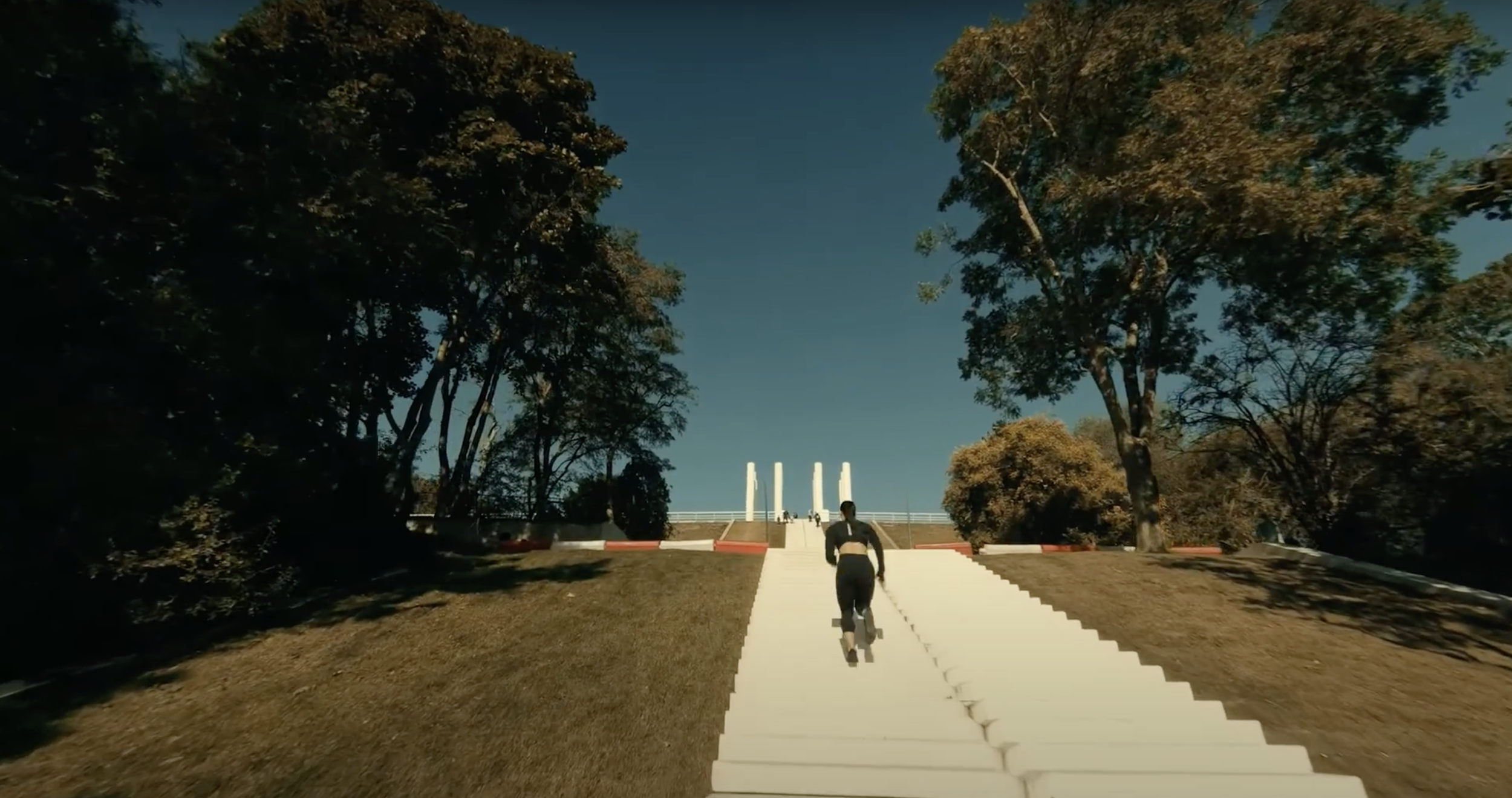 Une personne monte une grande rampe blanche bordée d'escaliers, entourée d'arbres et de ciel bleu.
