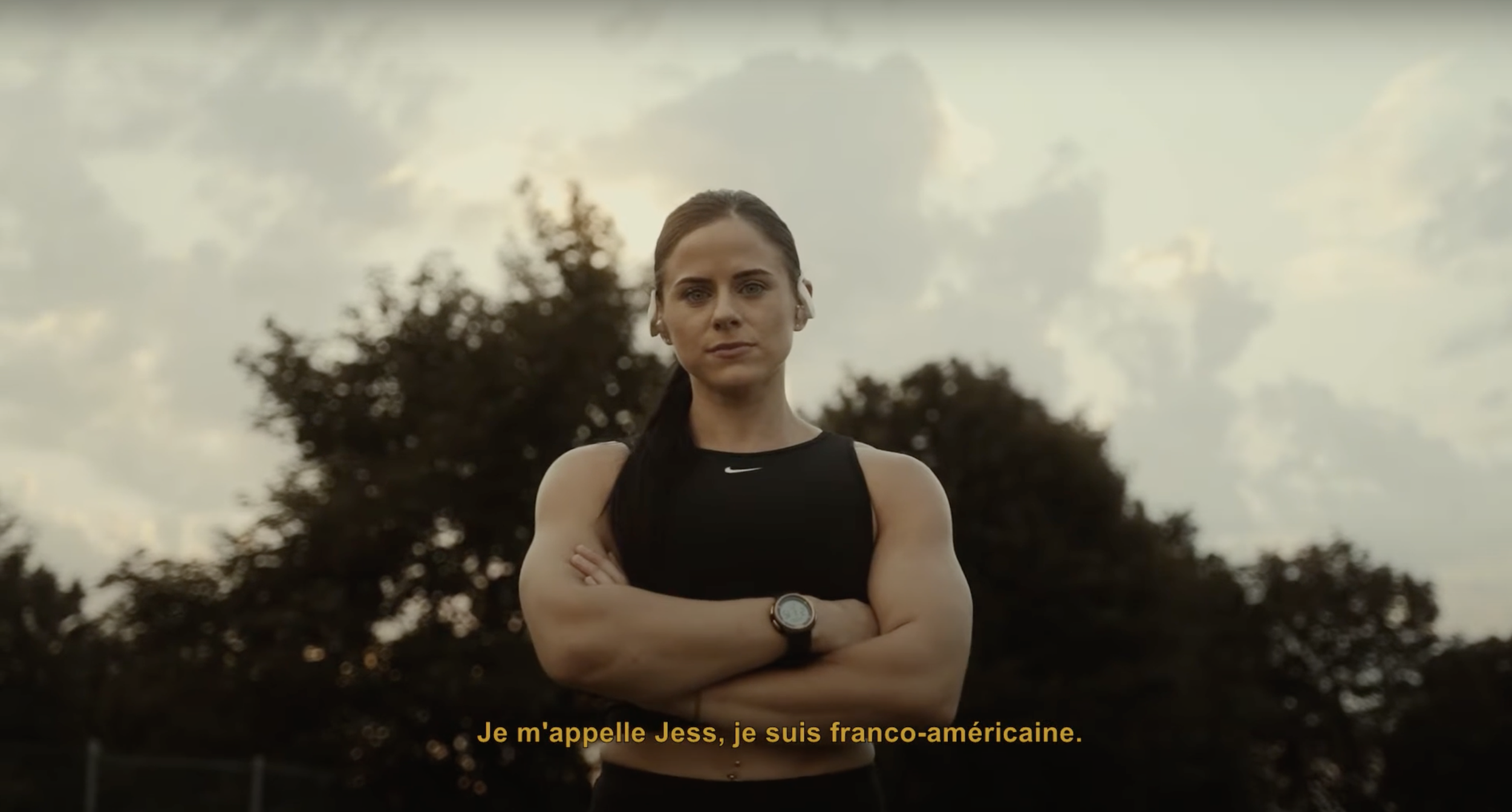 Une femme sportive se tient les bras croisés dehors avec un ciel nuageux en arrière-plan. Elle porte un débardeur noir, une montre et a les cheveux attachés. La légende en français indique qu'elle s'appelle Jess et qu'elle est franco-américaine.