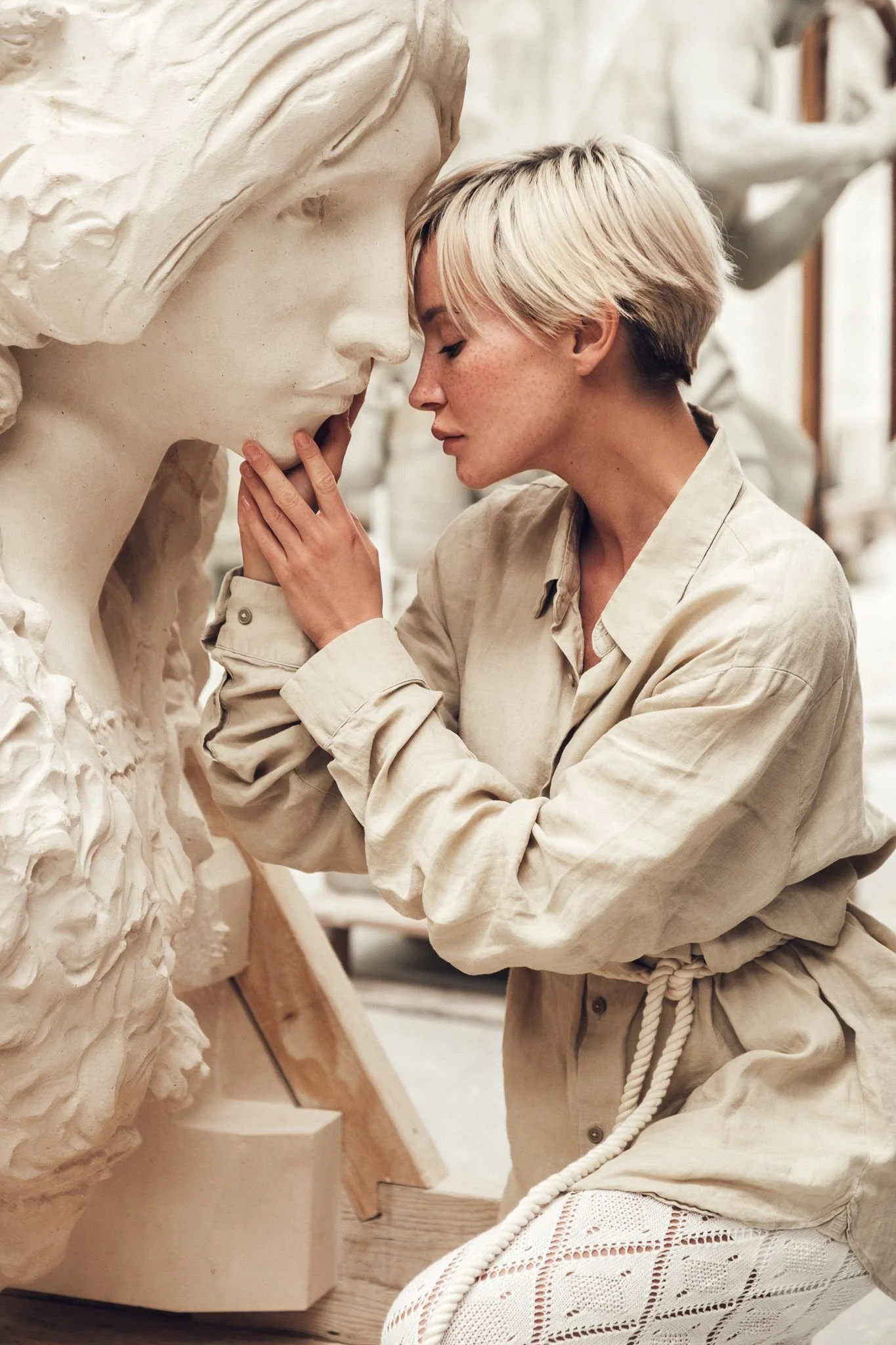 Une femme en atelier de sculpture, touchant une grande statue de femme en marbre, portant une tenue beige.