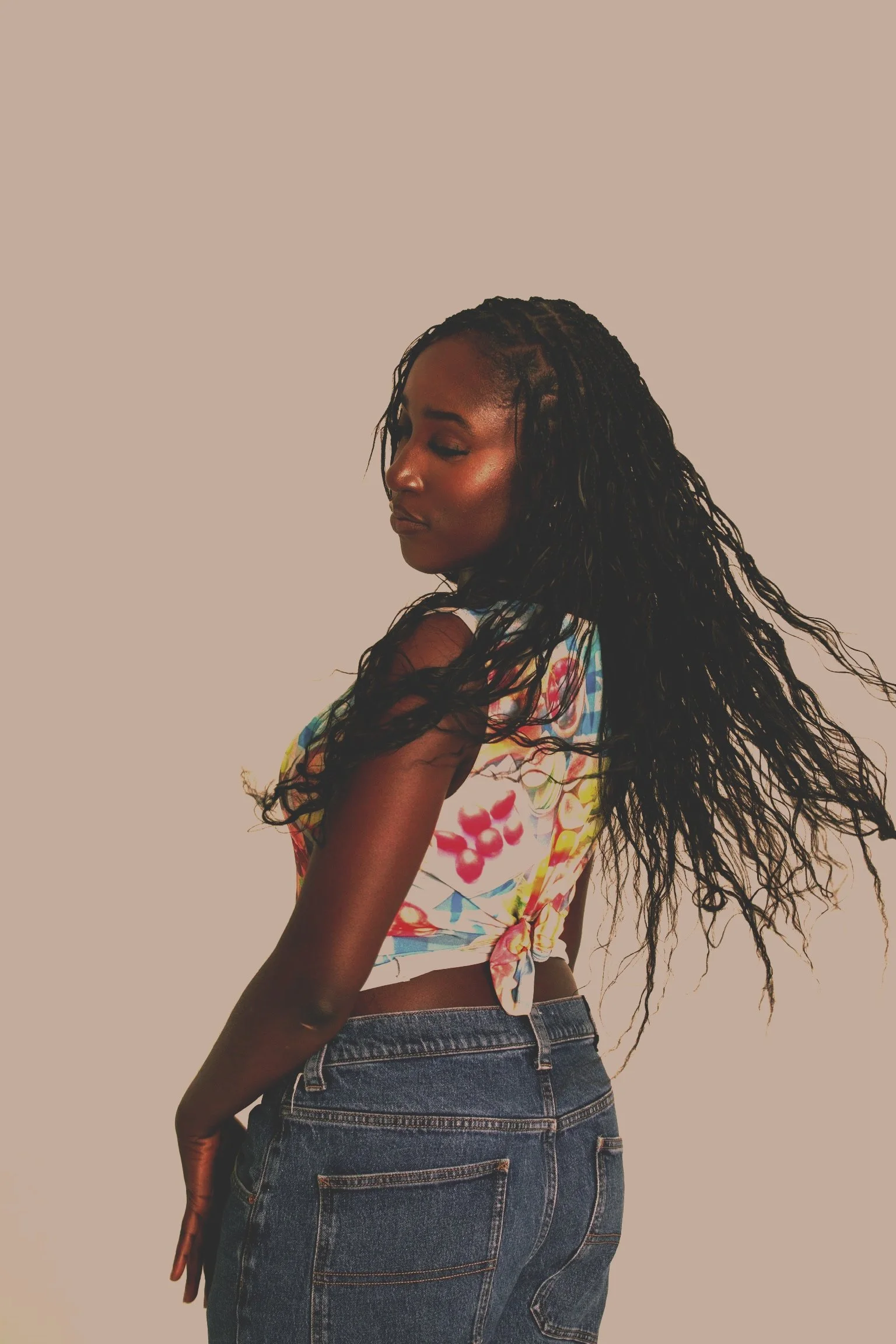 Young woman with long curly hair wearing a colorful floral top tied at the waist and denim jeans, standing against a plain light-colored background.