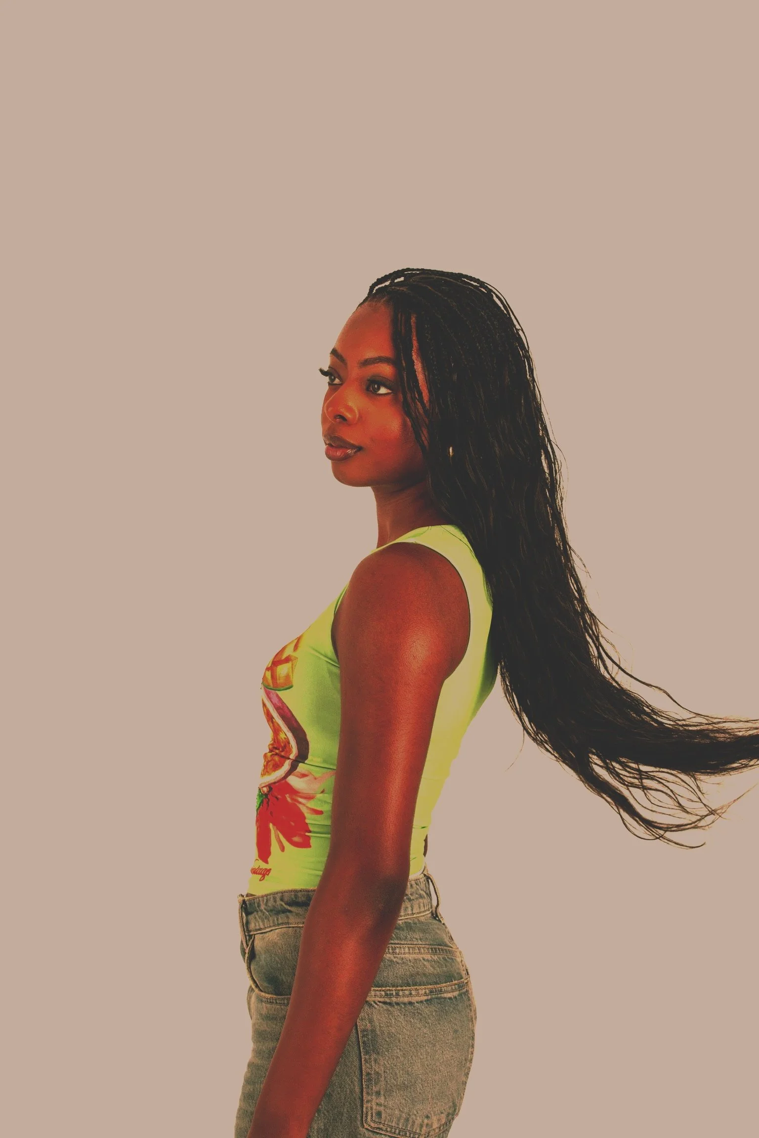 A woman with long, wavy black hair and a confident expression, wearing a colorful sleeveless top and high-waisted jeans, standing against a plain background.