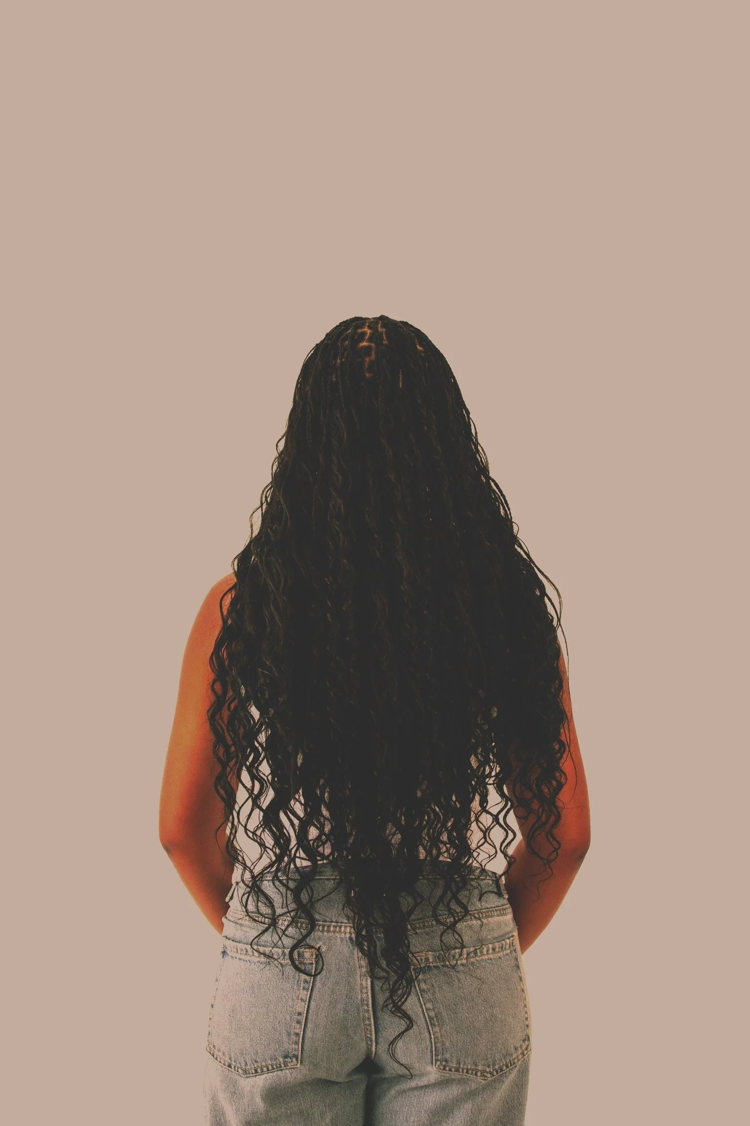 Back of woman with long, curly black hair wearing a sleeveless top and high-waisted jeans, standing against a plain background.