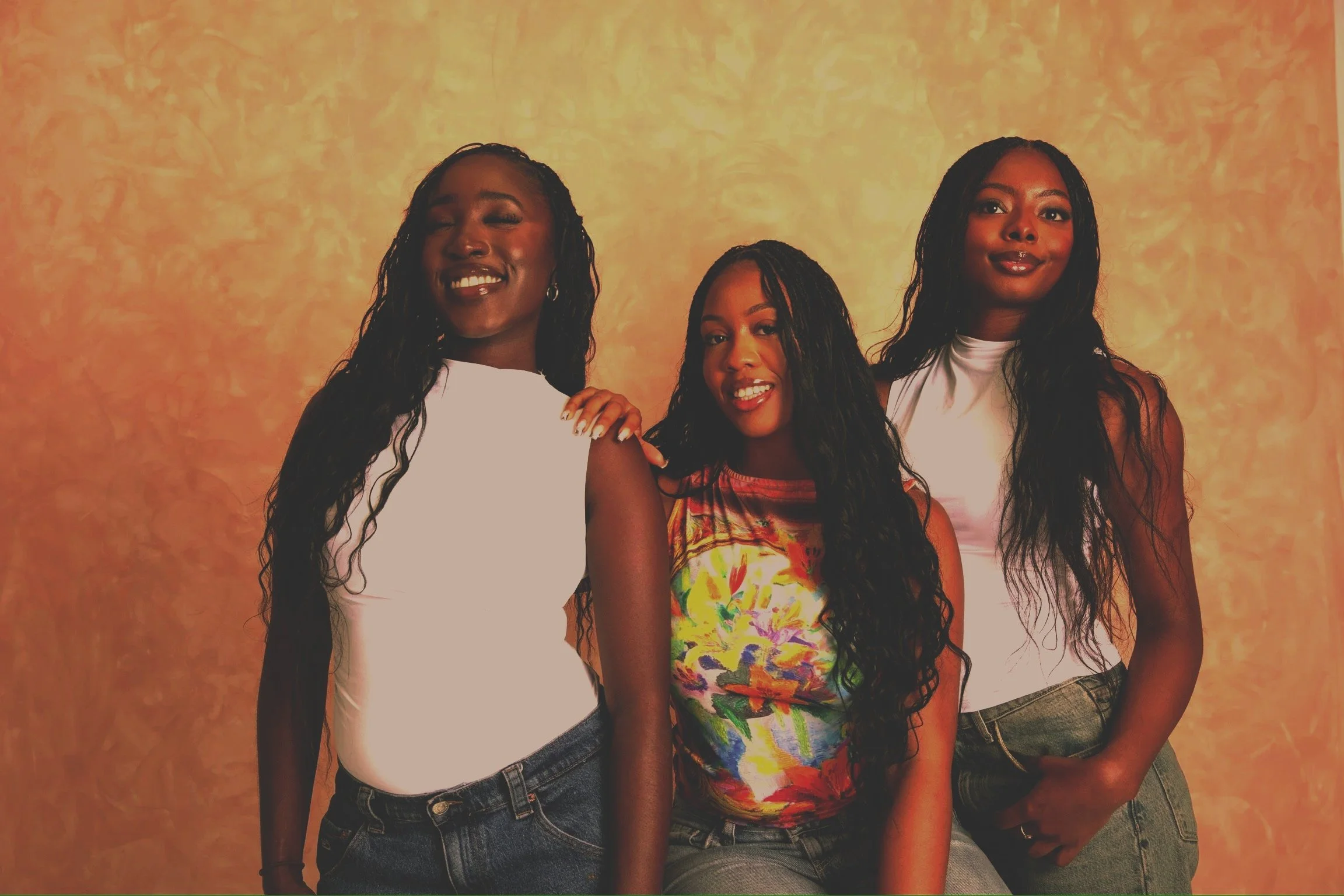 Three women with long, curly black hair standing together, smiling, against a textured beige background. The woman on the left wears a white sleeveless top and jeans, the woman in the center wears a colorful floral sleeveless top and jeans, and the woman on the right wears a white sleeveless top and gray jeans.