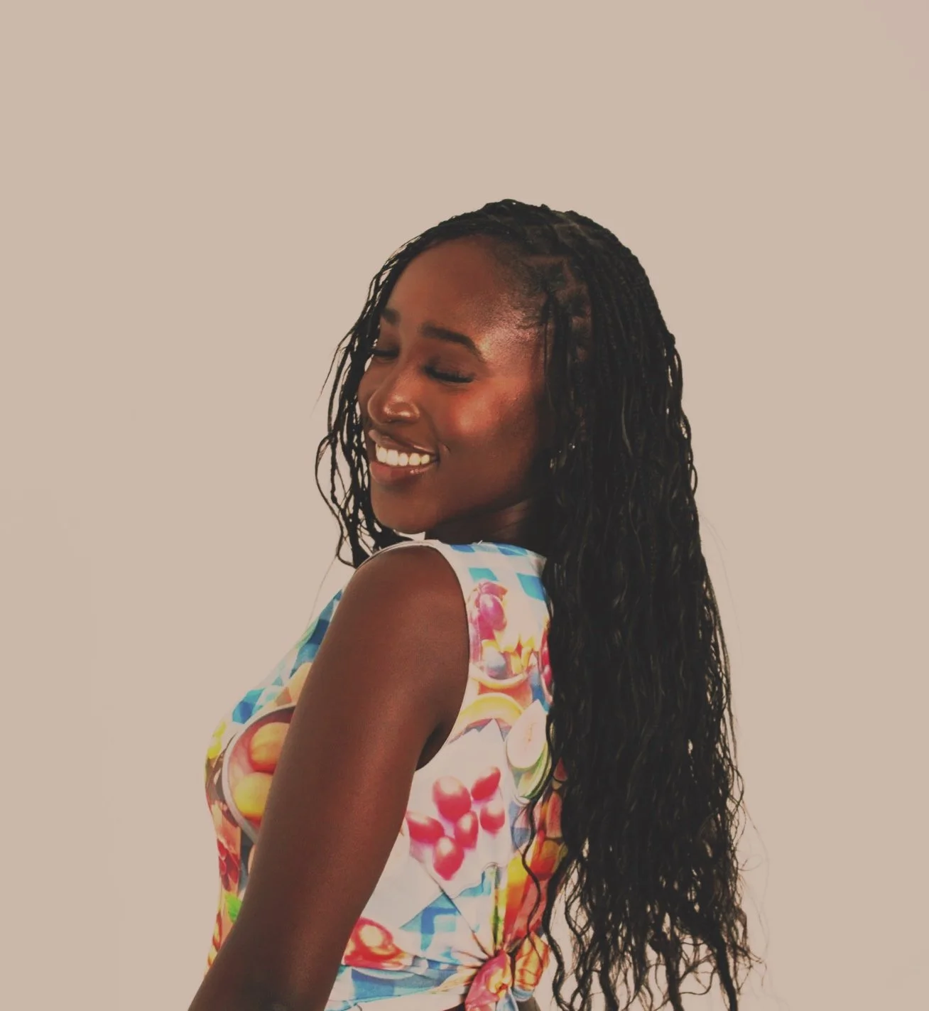 A young woman with long, curly black hair, smiling with her eyes closed, wearing a colorful floral sleeveless top, against a plain beige background.