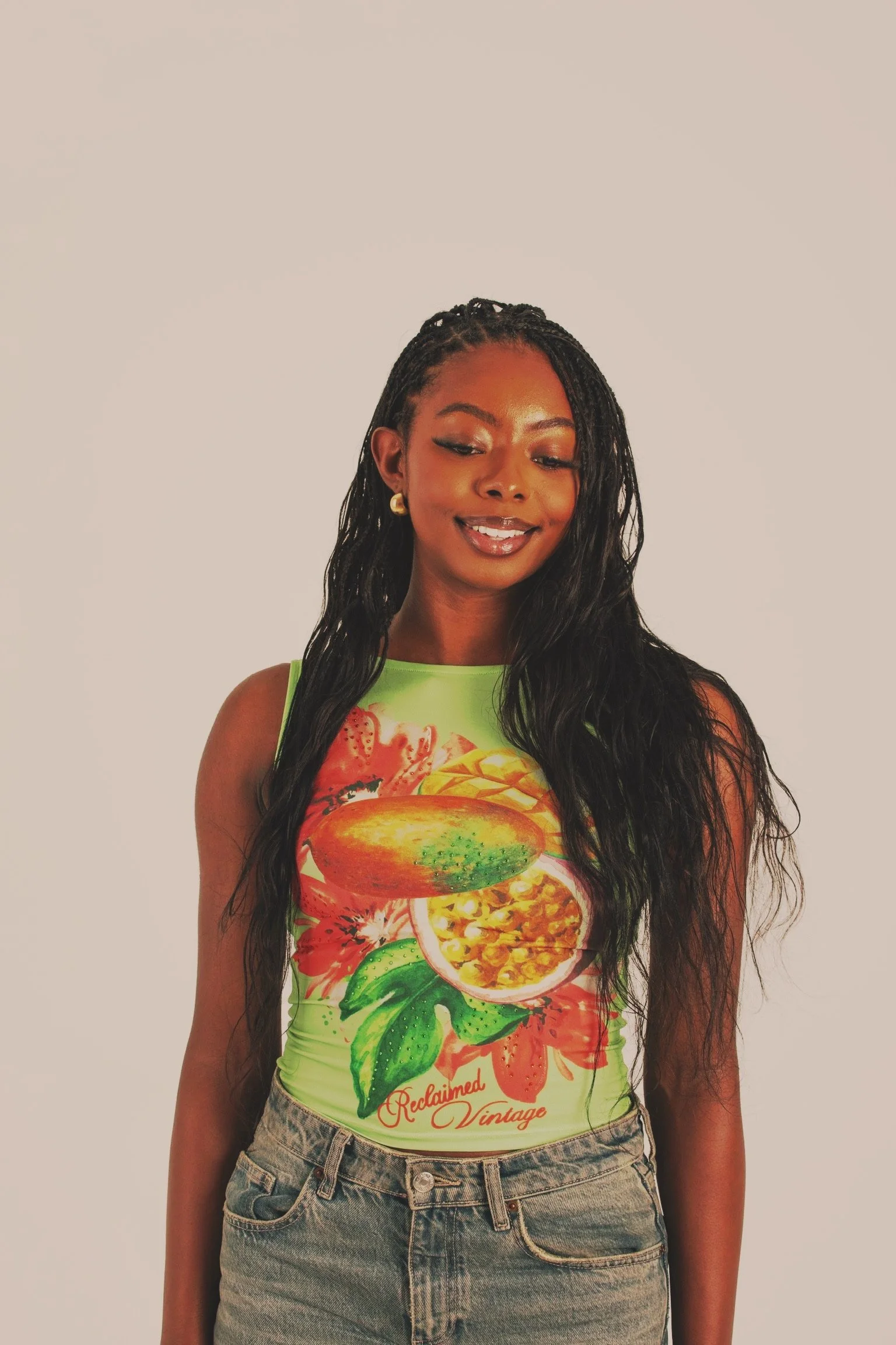 A young woman with long, wavy black hair, wearing a sleeveless neon green top with fruit and floral graphics and the words 'Reclaimed Vintage,' standing against a plain light background.