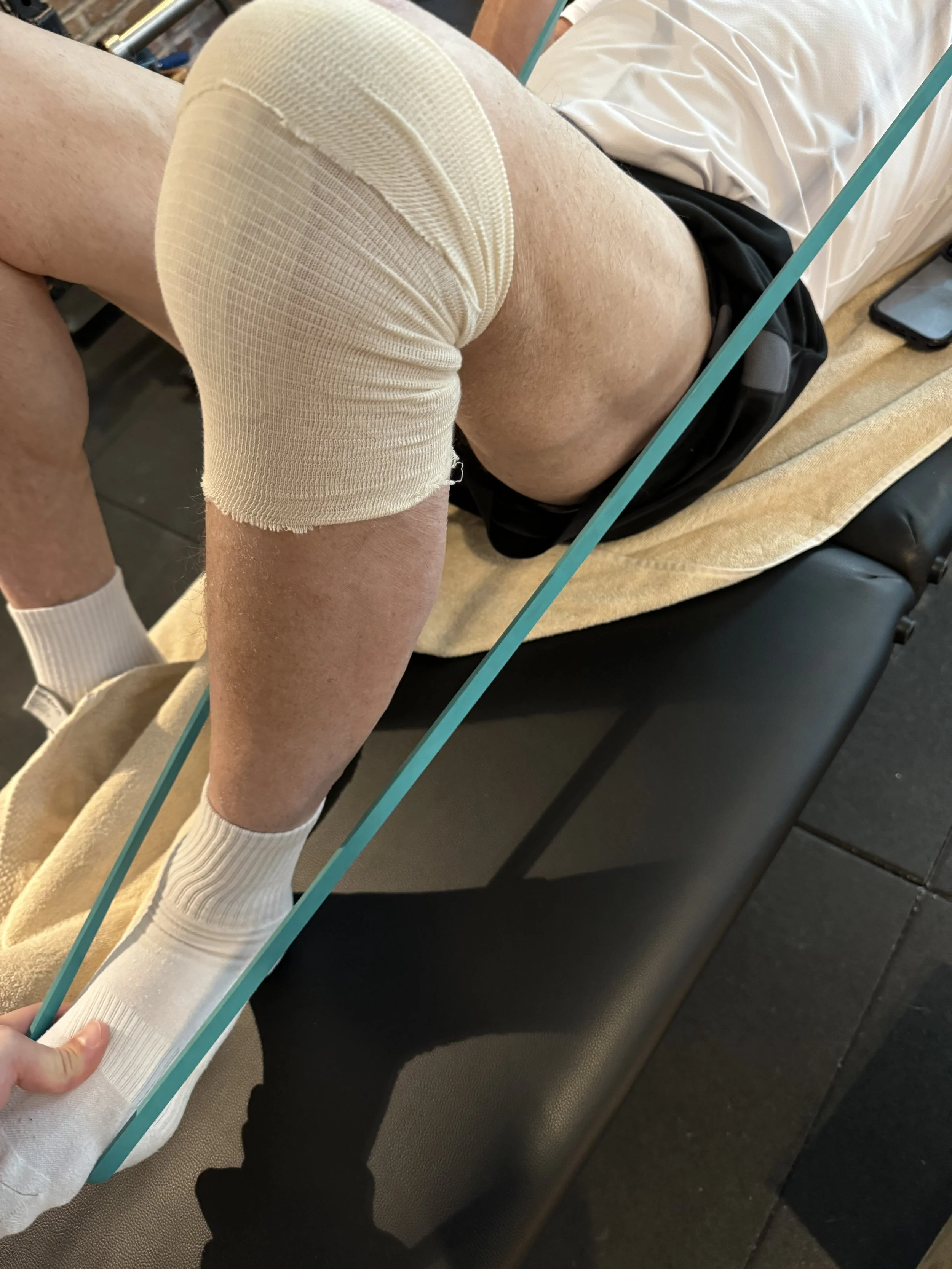 Paciente operado de prótesis de rodilla recibe rehabilitación con ejercicio terapéutico usando gomas elásticas.