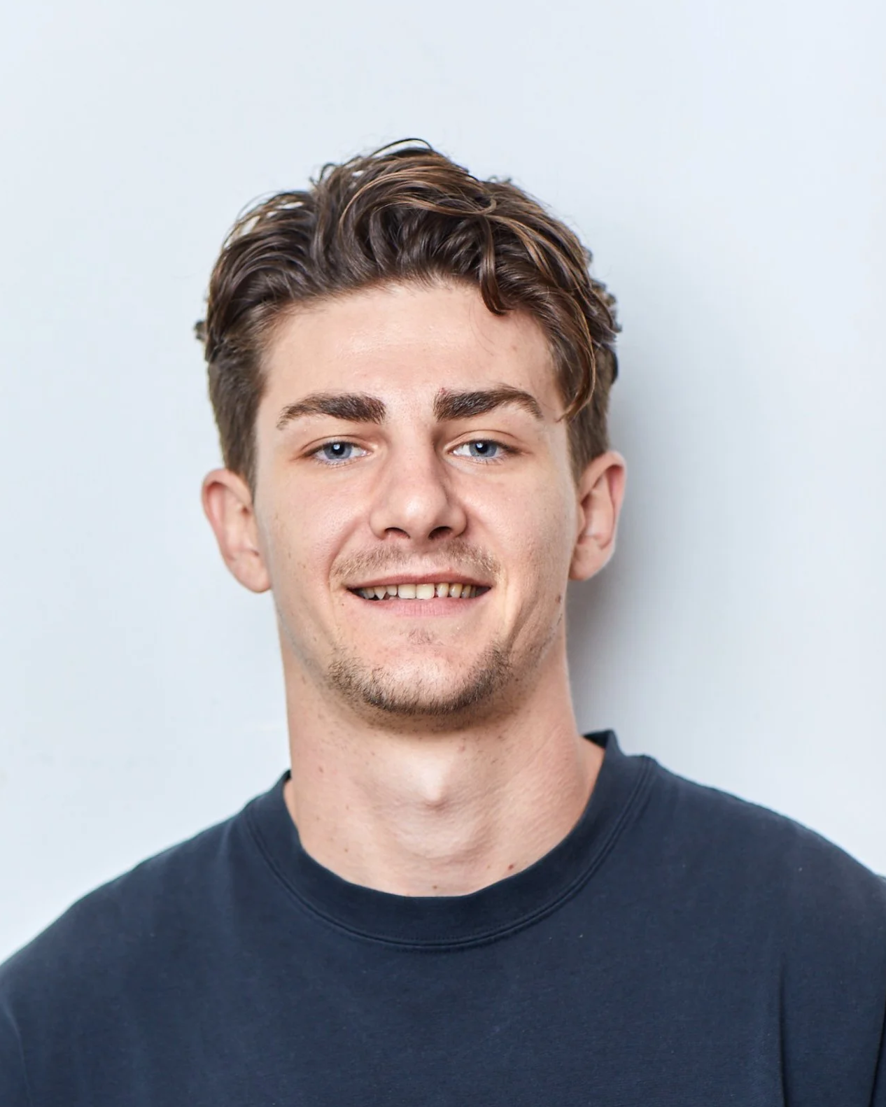 Retrato de un joven con cabello castaño y ojos azules, sonriendo ligeramente, vistiendo una camiseta negra, contra fondo blanco.