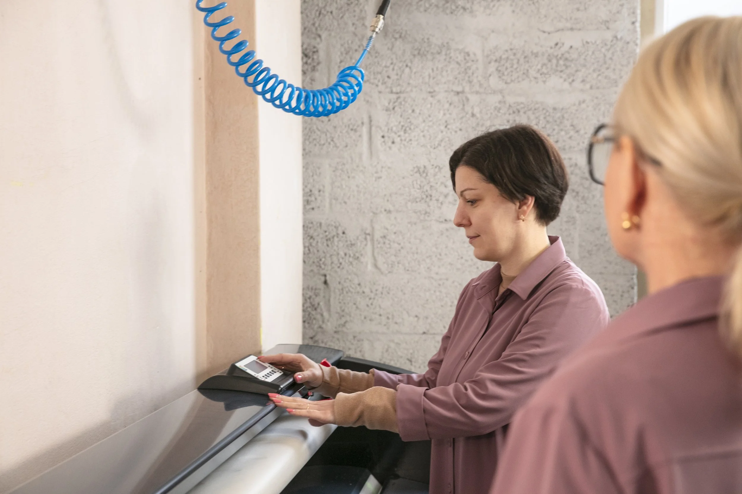Two women work on a manufacturing assembly line. The woman in focus has short dark hair and wears a mauve shirt, while the other woman has blond hair tied back, wears glasses and a similar shirt. They are handling a small electronic device on a conve