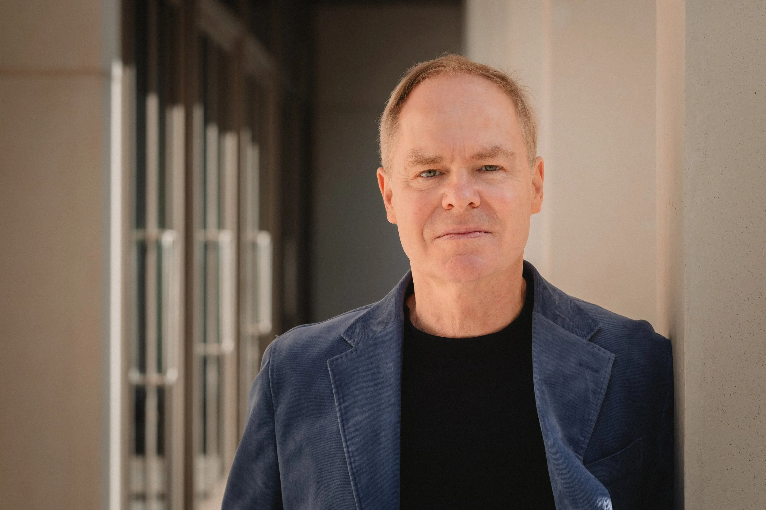 A man with blond hair and blue eyes standing outdoors next to a beige wall, wearing a dark jacket over a black shirt.