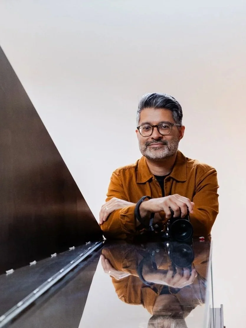 A man with glasses and a salt-and-pepper beard, wearing a brown jacket, sitting at a reflective black surface with a background of plain, light-colored wall. He has a camera in his right hand resting on the surface and is smiling at the camera.