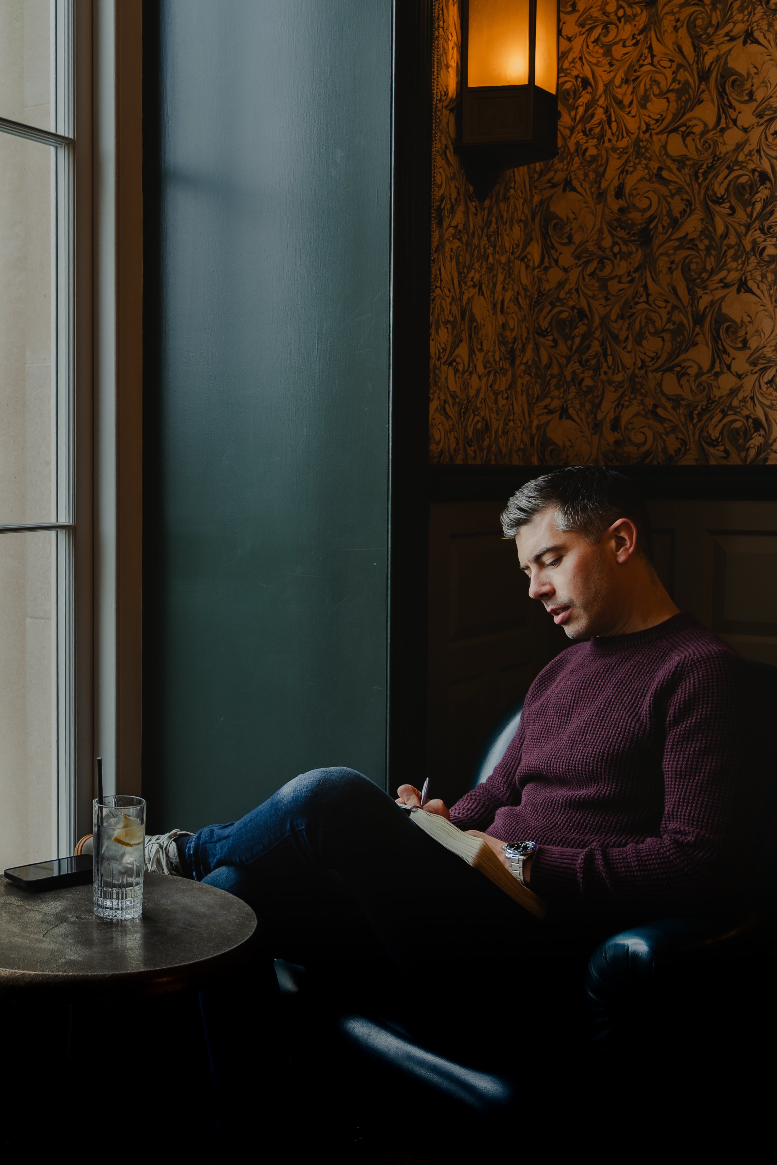 A man sitting by a window in a cafe, writing in a notebook with a pen, next to a glass of water and a smartphone on the table.
