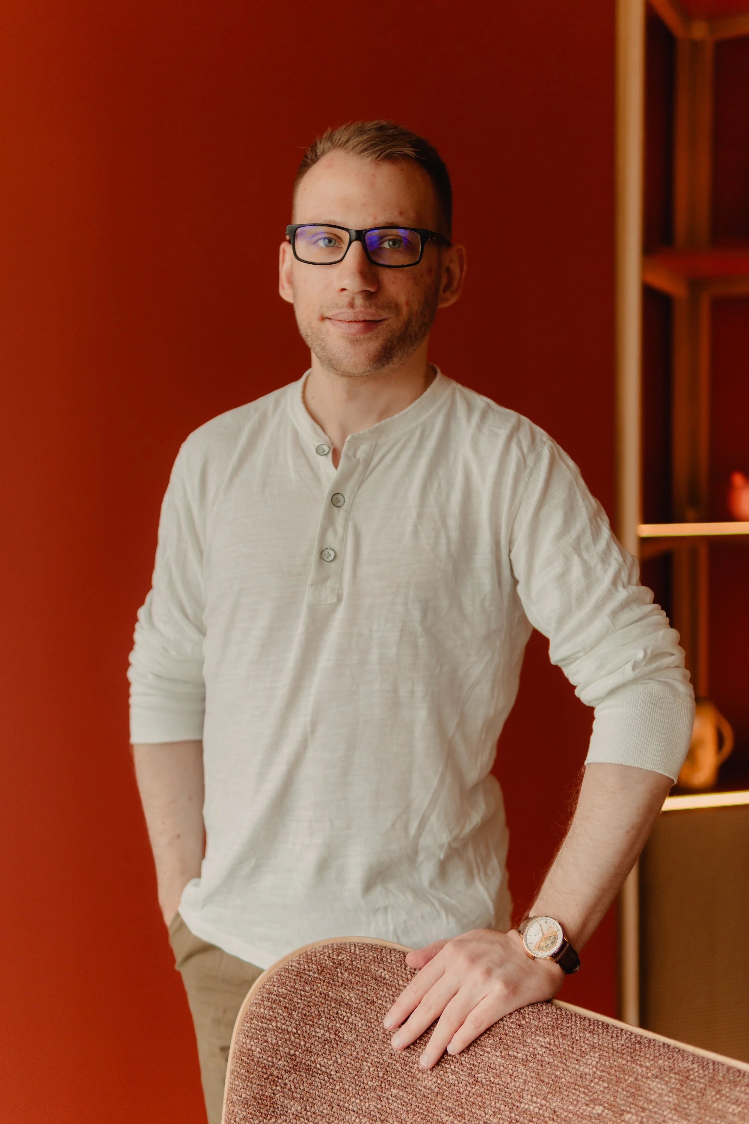 A man with short hair, glasses, and a smartwatch, standing in front of a red wall with his hand resting on a chair.
