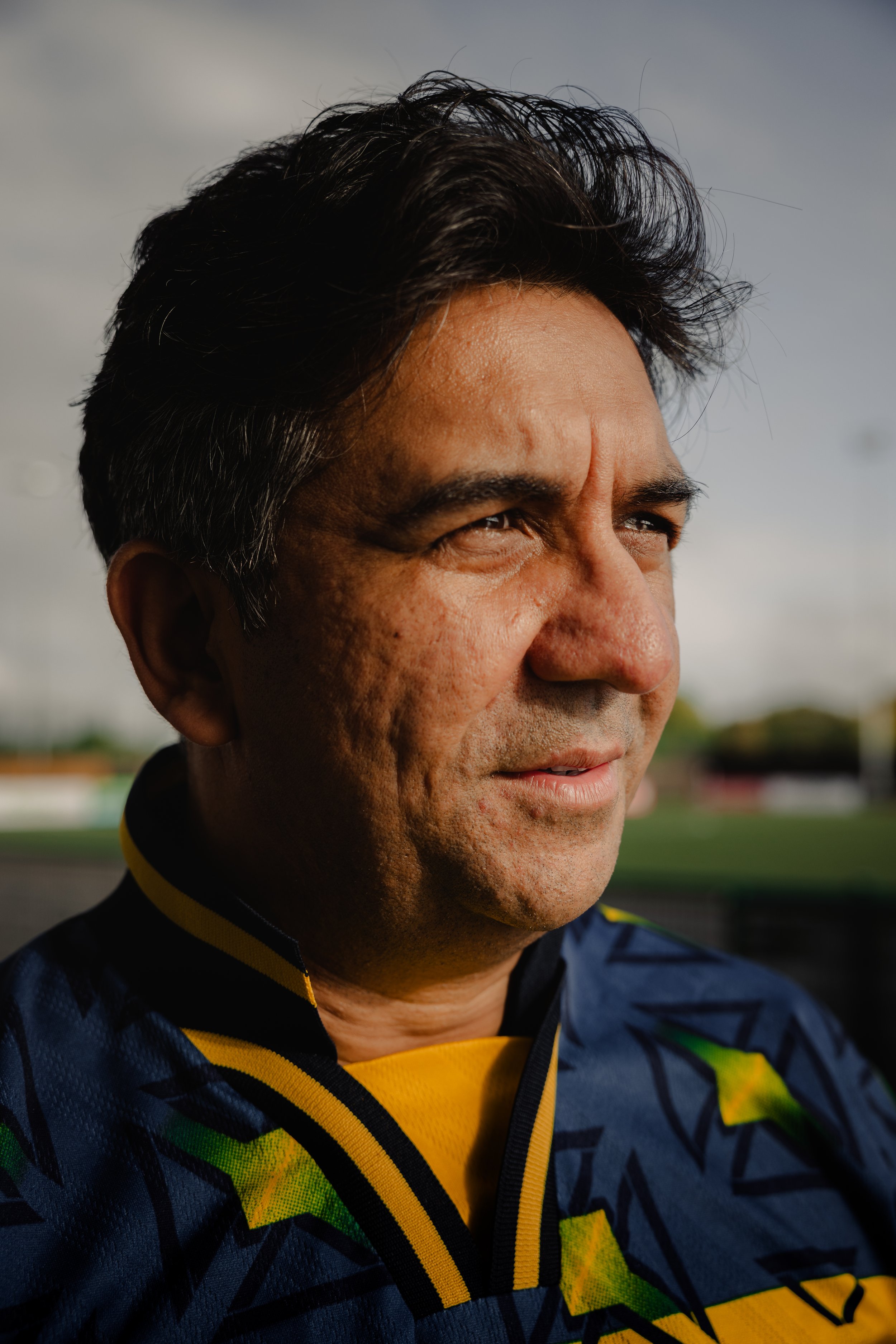 Close-up of a middle-aged Indian man with dark hair, looking thoughtfully into the distance, wearing a sports jacket with yellow and green accents, outdoors on a cloudy day.