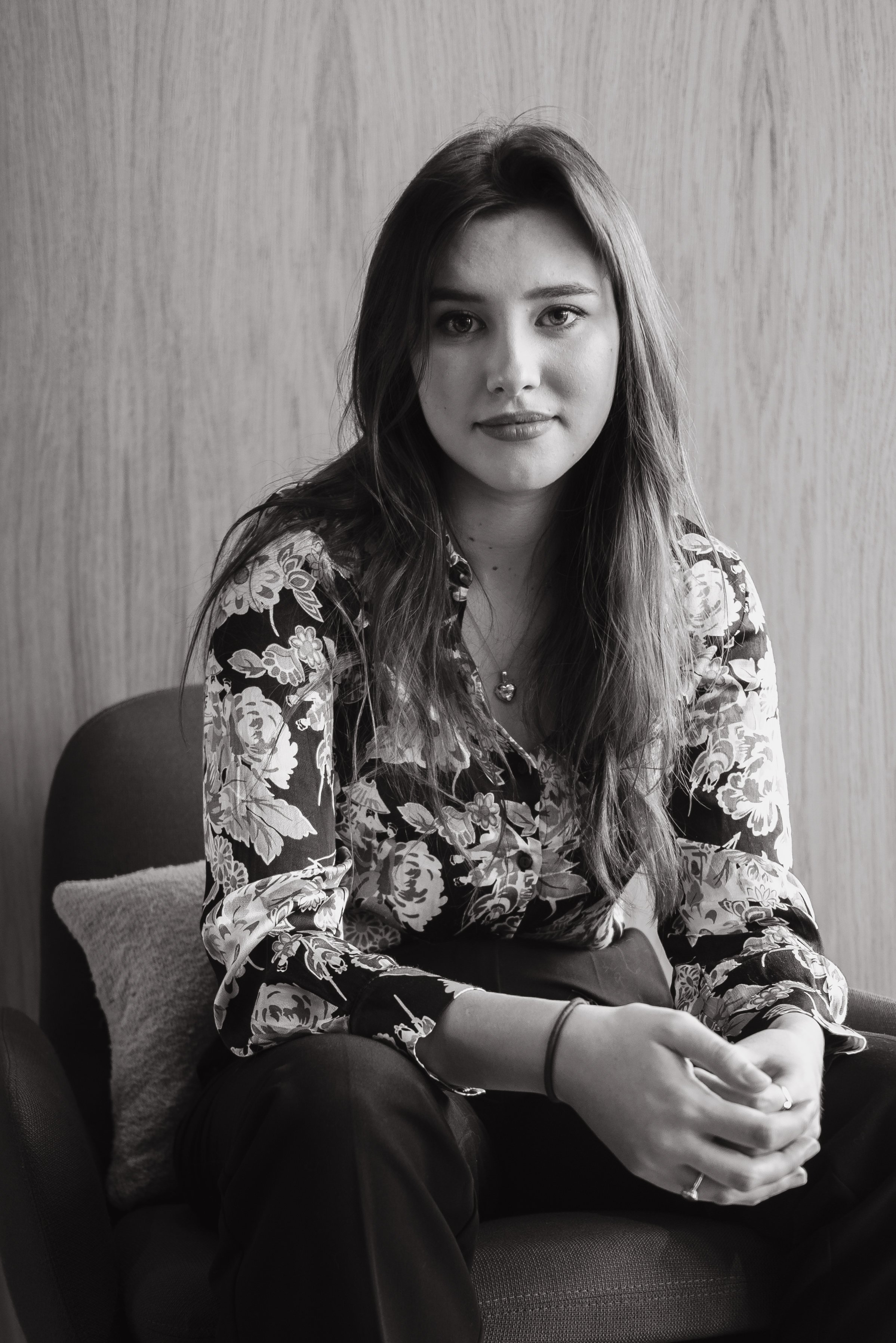 A young woman with long hair sitting on a chair, looking at the camera, wearing a floral patterned long sleeve shirt, in front of a wooden wall.