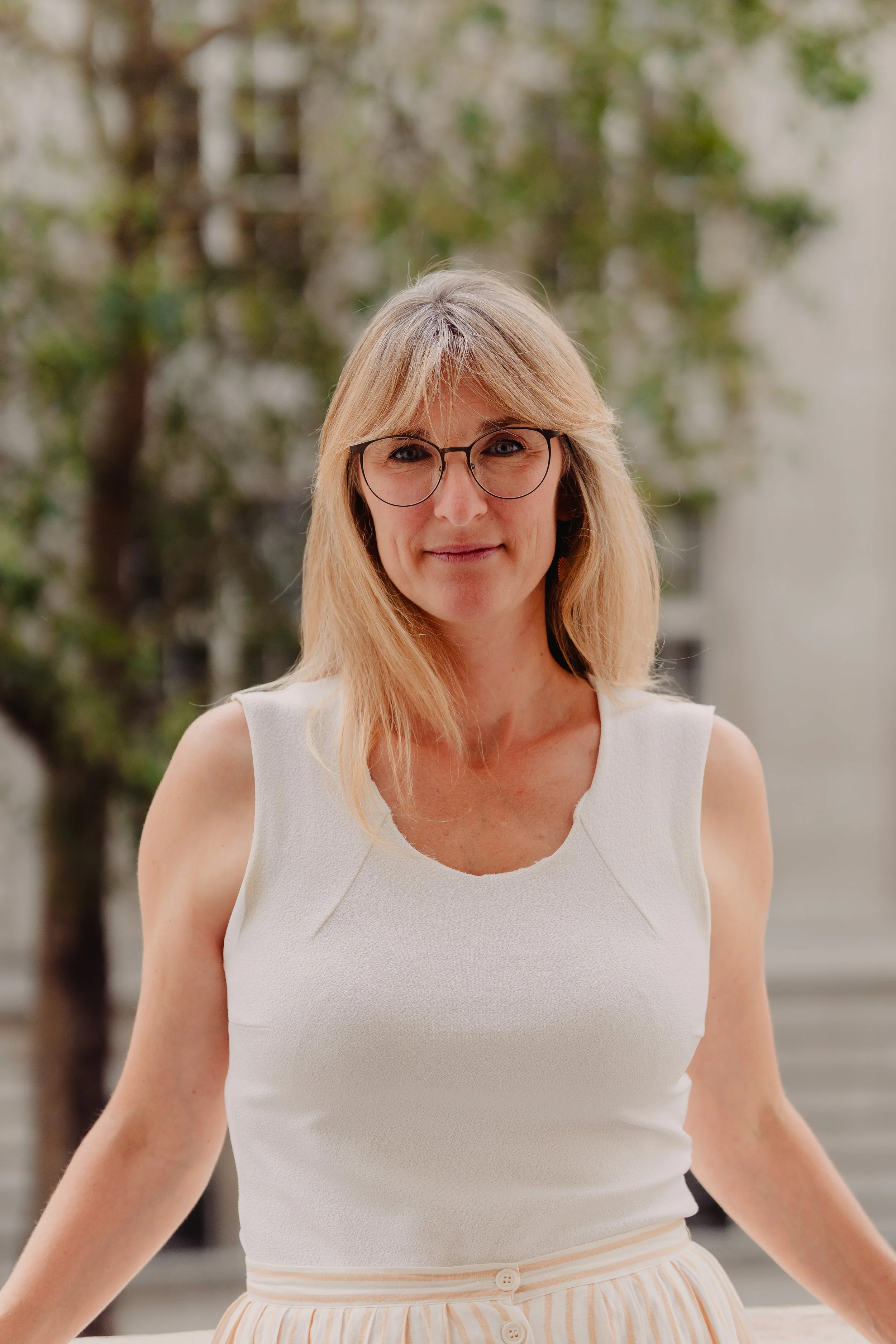 A woman with blonde hair wearing glasses and a sleeveless white top, standing outdoors with greenery in the background.