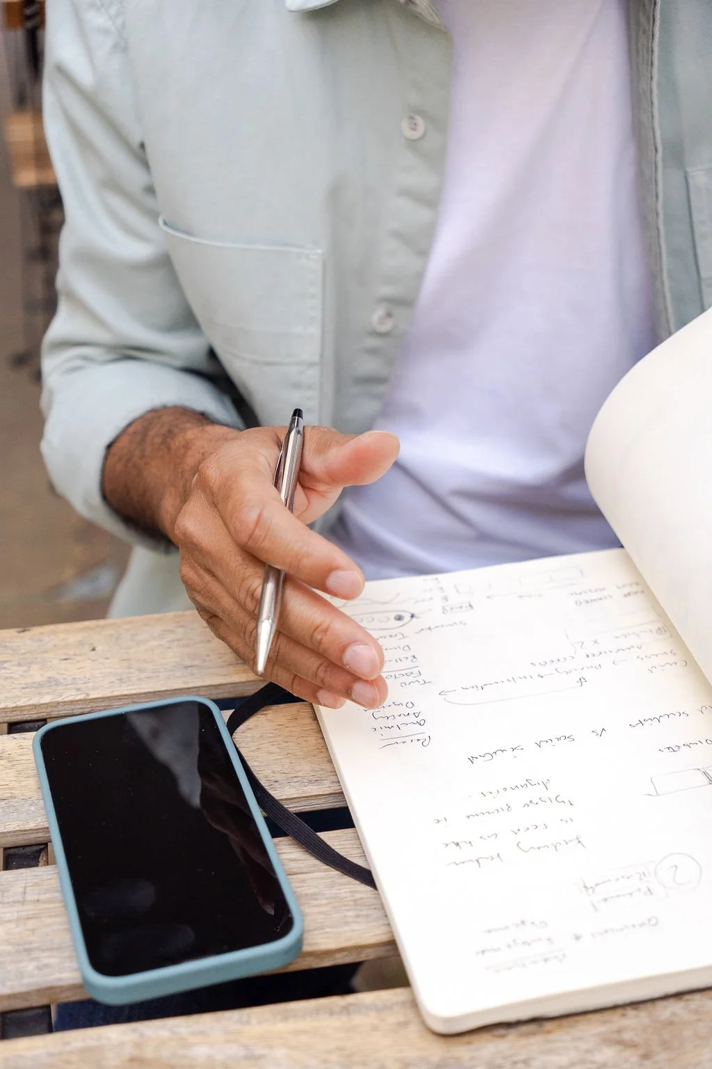 Close-up of a person writing notes with a pen in a notebook, with a smartphone resting on a wooden table.