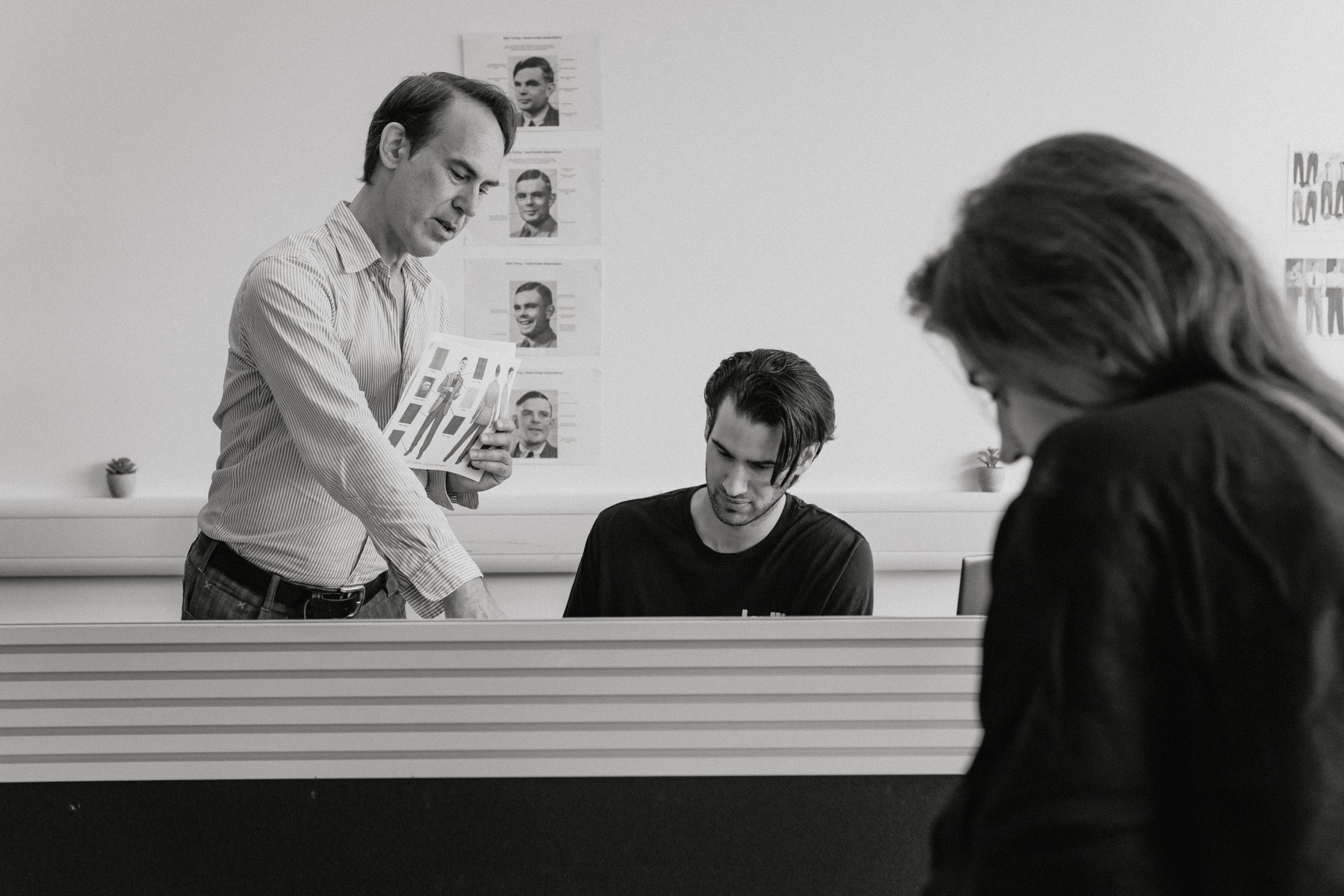 Scene inside an office with three people. A man standing and holding a brochure or report, another listening and a woman in the foreground looking down. The background features framed photos or posters on the wall.