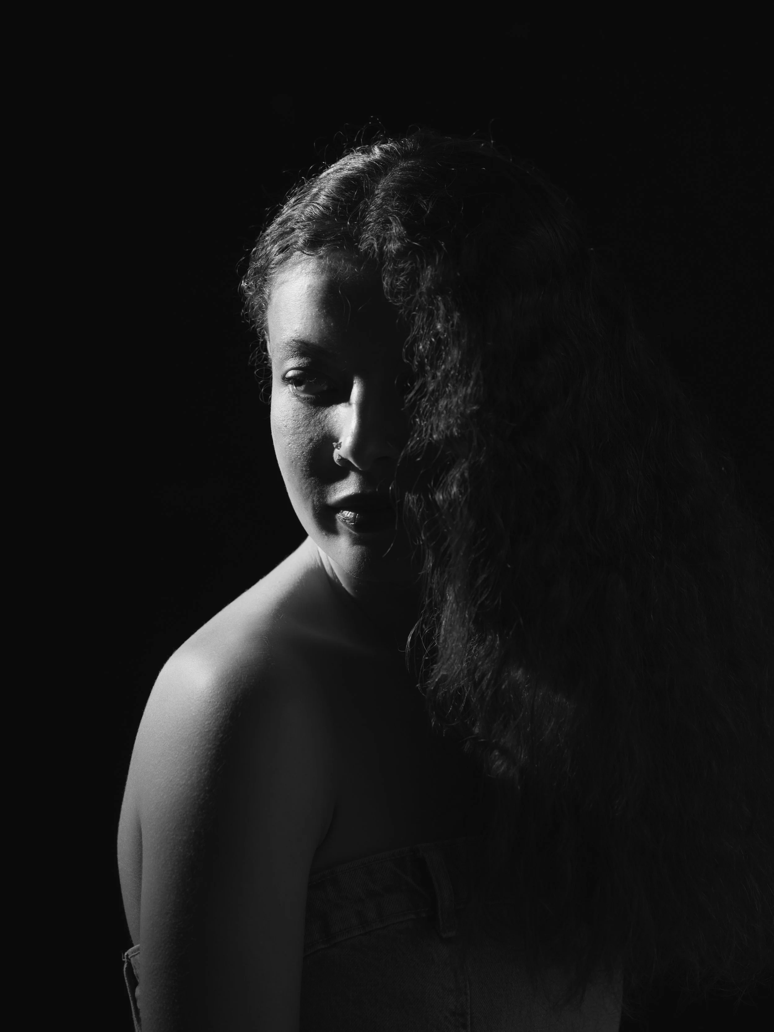 A black and white portrait of a woman with one side of her face and shoulder visible. She has curly hair covering part of her face and is wearing a strapless top.
