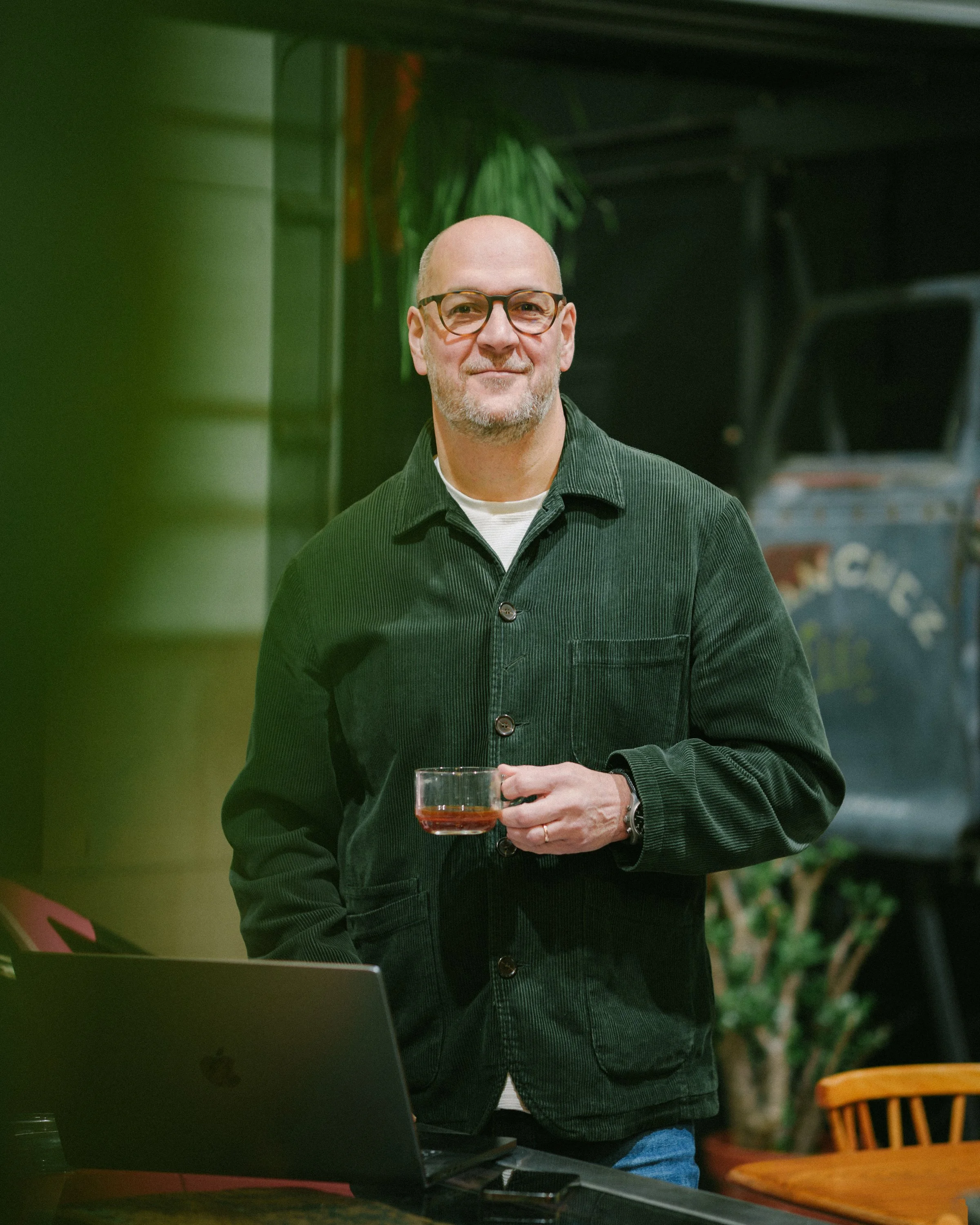A bald man wearing glasses and a dark green button-up shirt holding a drink, standing in a room with green and black walls and a wooden chair.