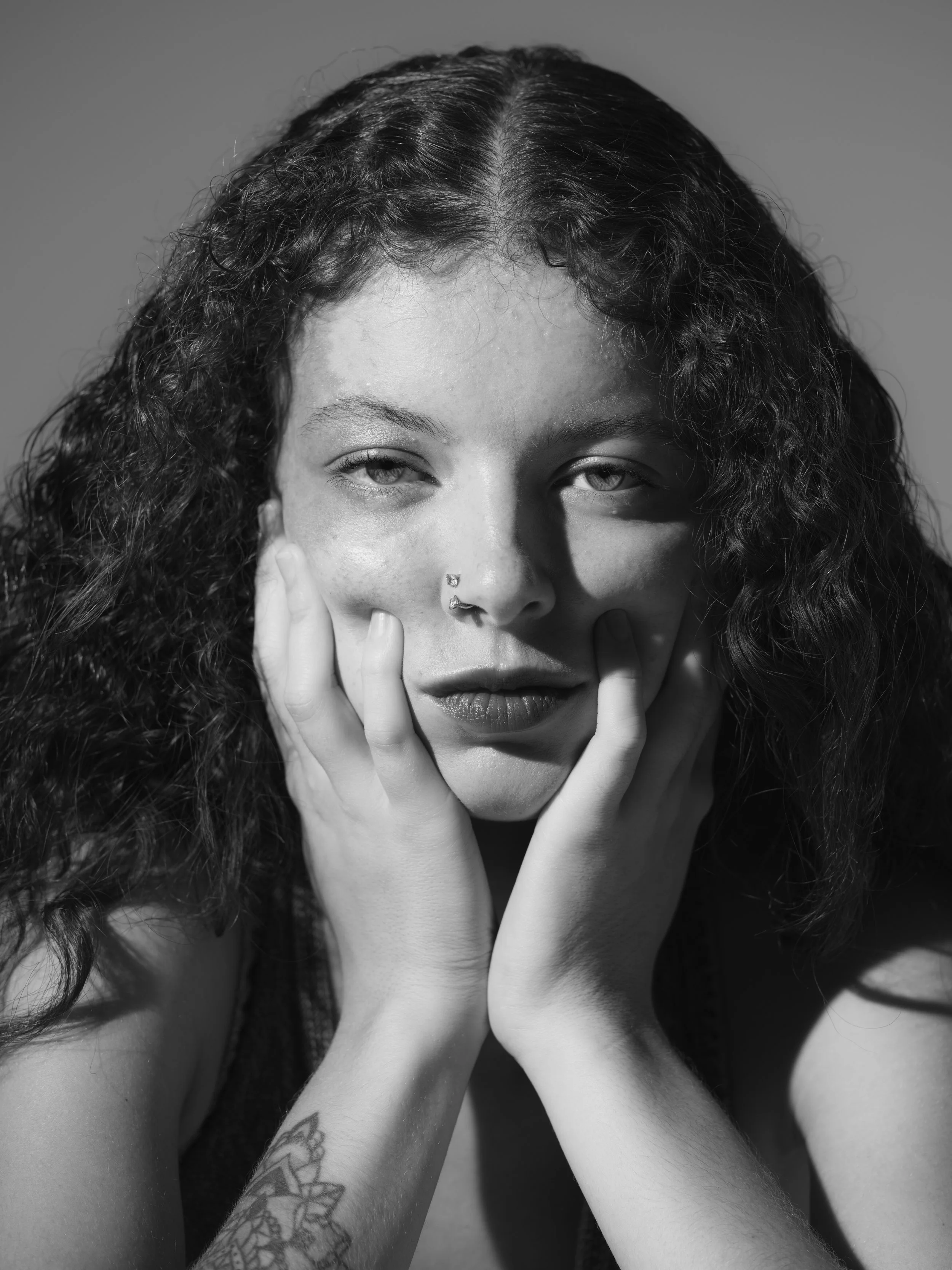 Black and white photo of a woman with curly hair, one eye closed, resting her face on her hands, with a nose ring and visible tattoo on her arm.