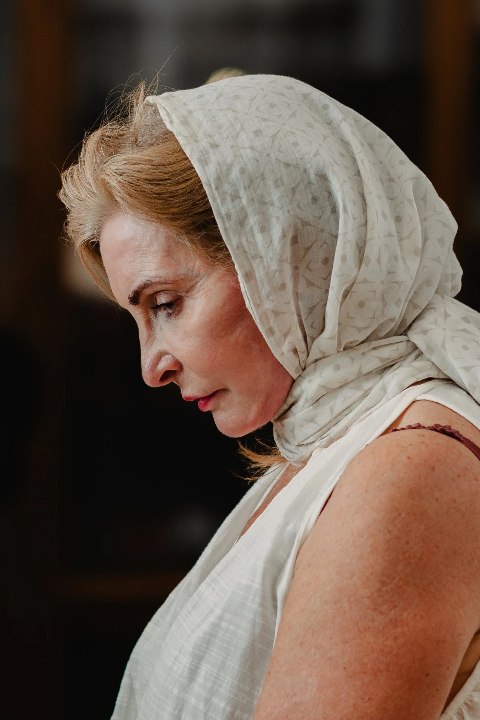 A woman with light skin and red hair, wearing a light-colored headscarf and sleeveless top, looking down.