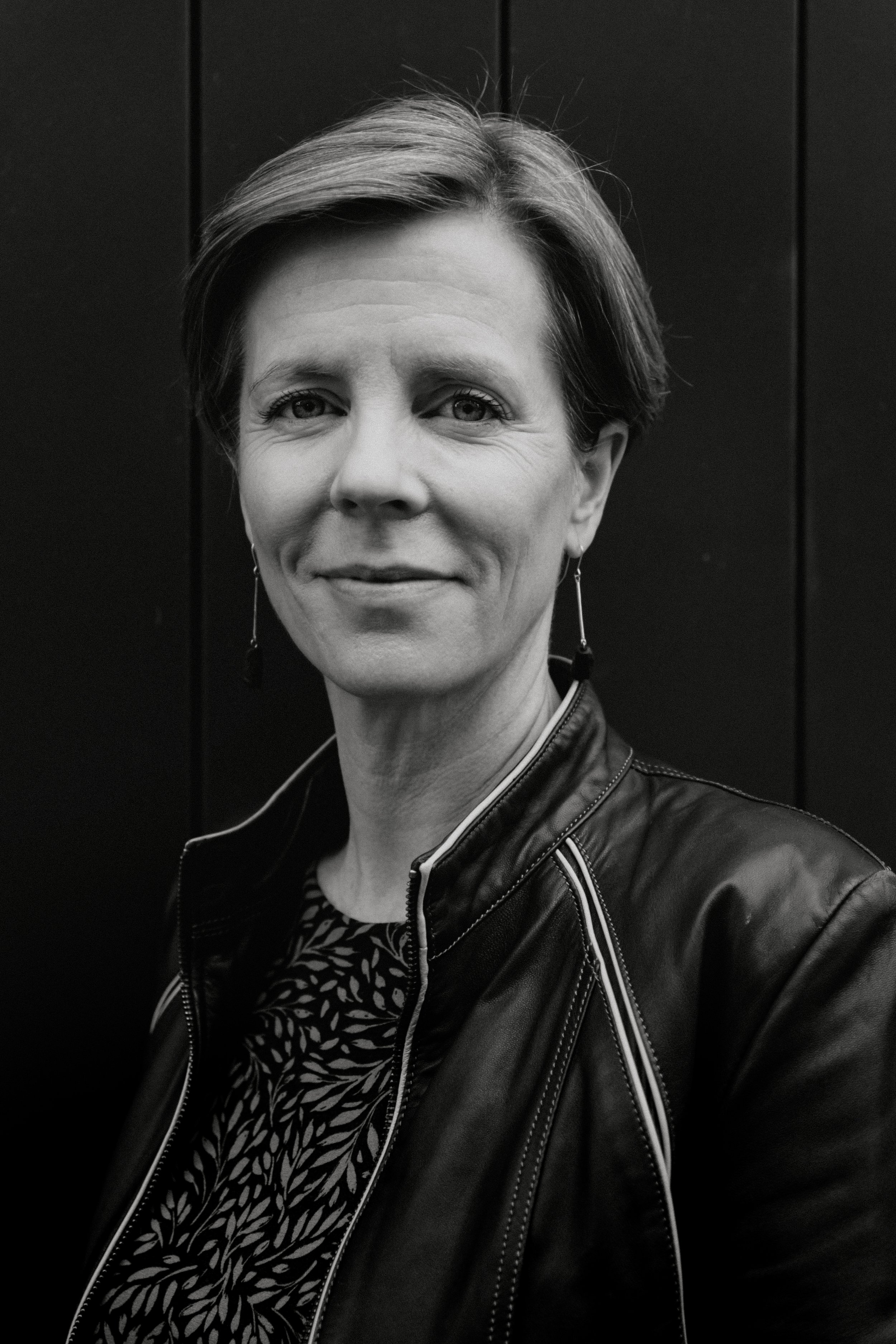 A woman with short hair wearing a leather jacket and patterned top, standing in front of a dark wall.