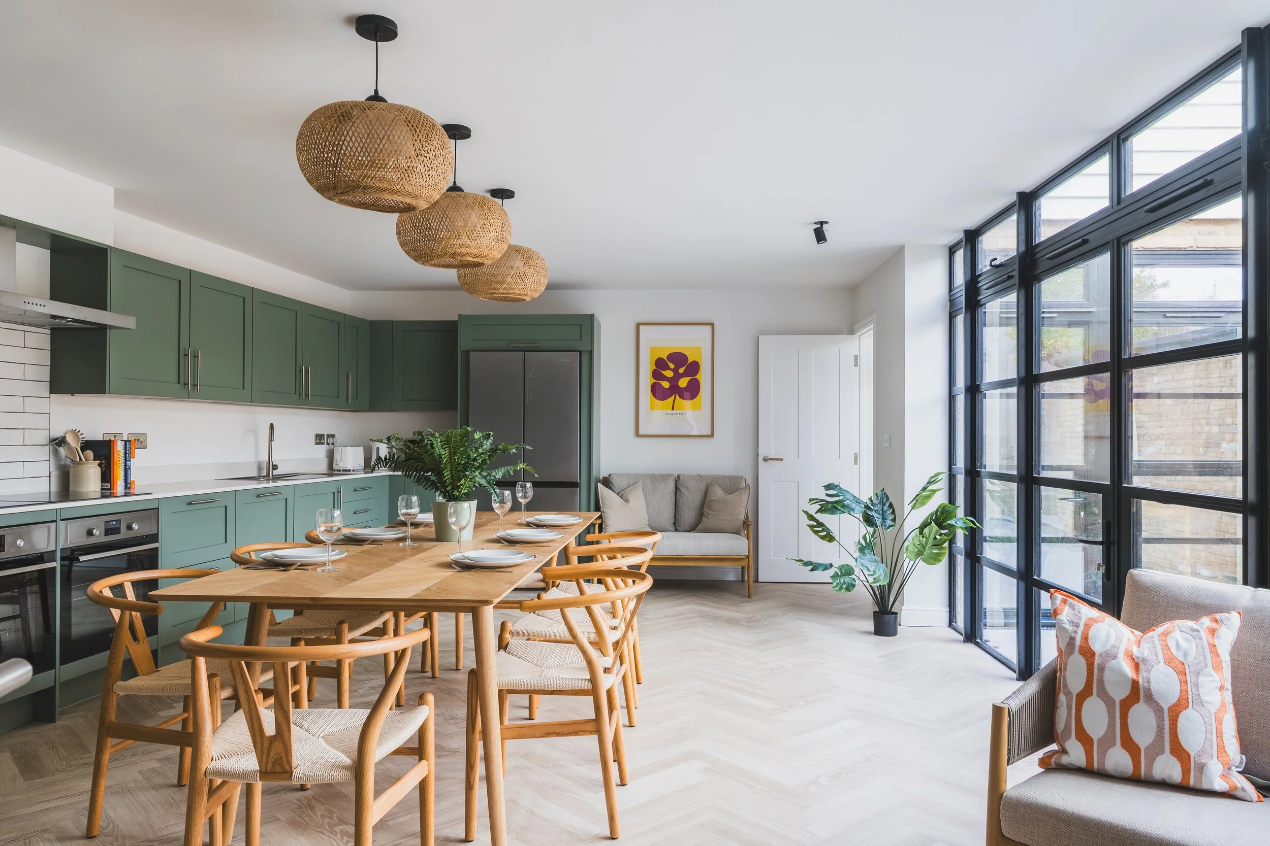 Modern open-concept kitchen and dining area with green cabinets, wooden table, woven pendant lights, large windows, and indoor plants.
