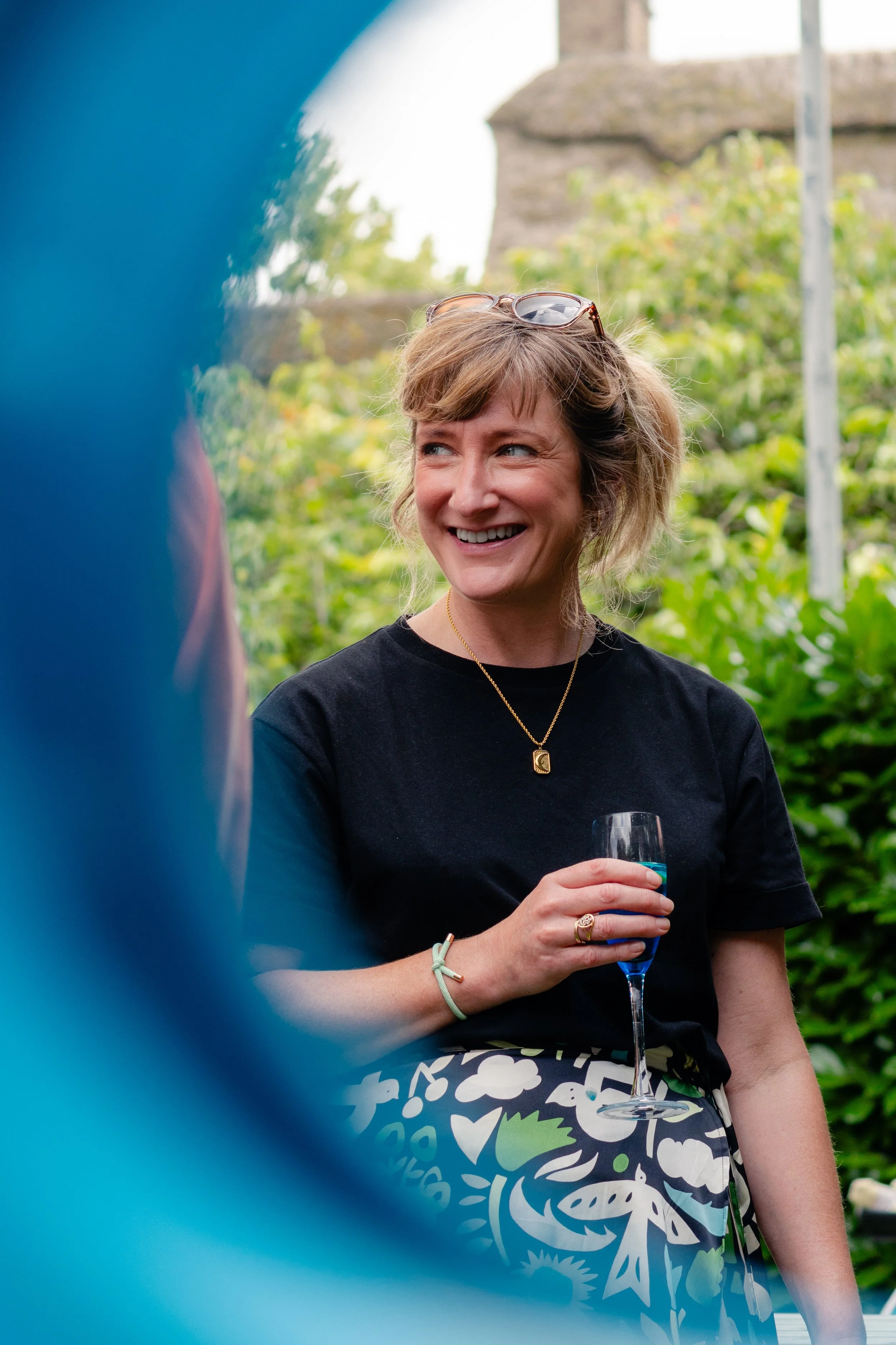 A woman with short blonde hair, wearing sunglasses on her head, smiling and holding a glass of blue drink at an outdoor gathering. She is dressed in a black top and a patterned skirt, with a forested background.
