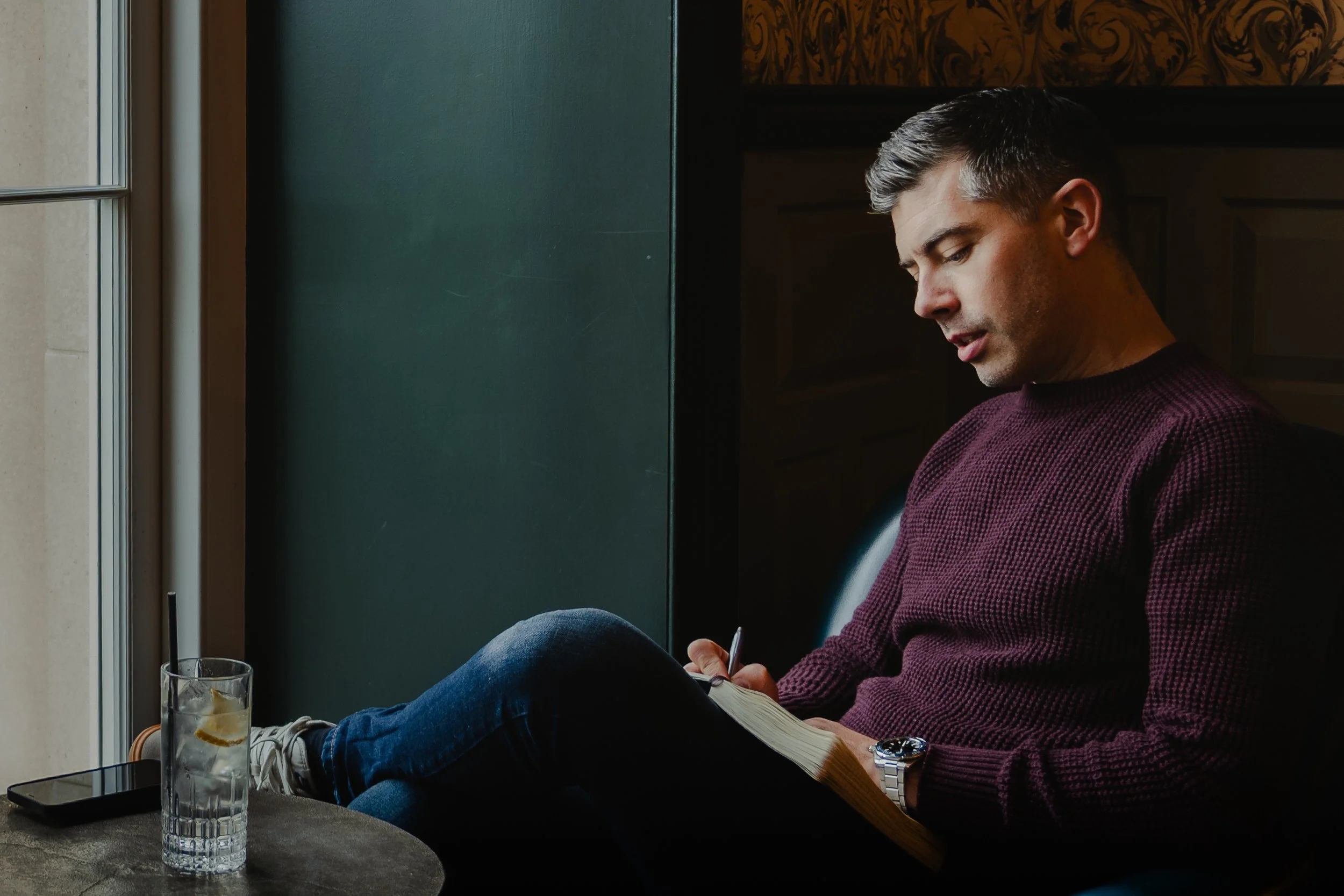 A man sitting in a cozy cafe, writing in a notebook with a pen, with a glass of water and a smartphone on the table beside him.