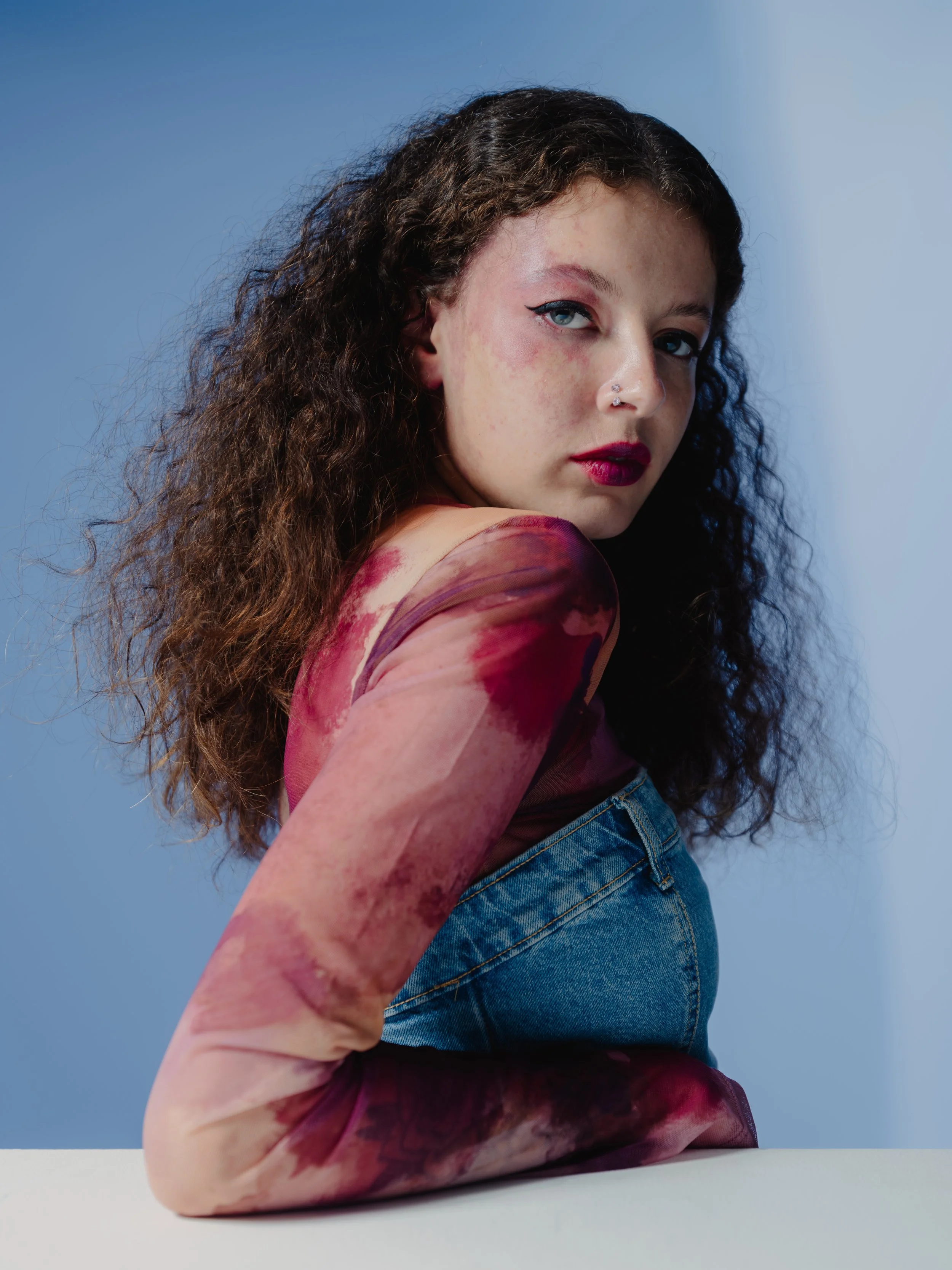 A young woman with curly brown hair and fair skin, wearing a sheer, patterned dress with pink, purple, and beige tones, and blue jeans, sitting against a blue background, looking at the camera.