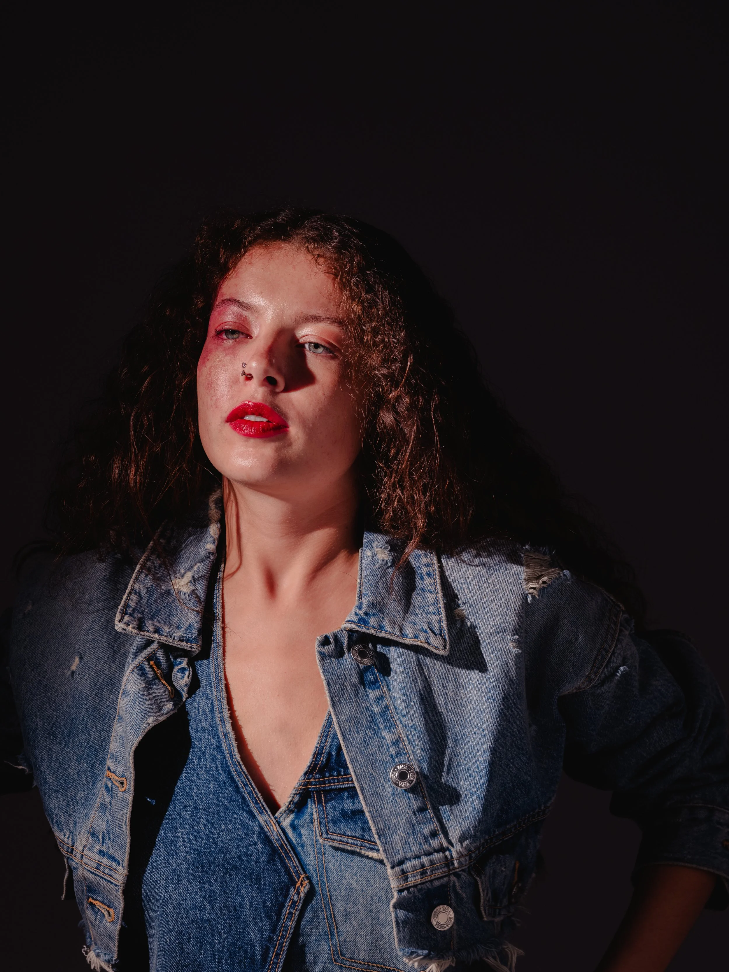 Portrait of a woman with dark curly hair wearing a distressed denim jacket against a dark background.