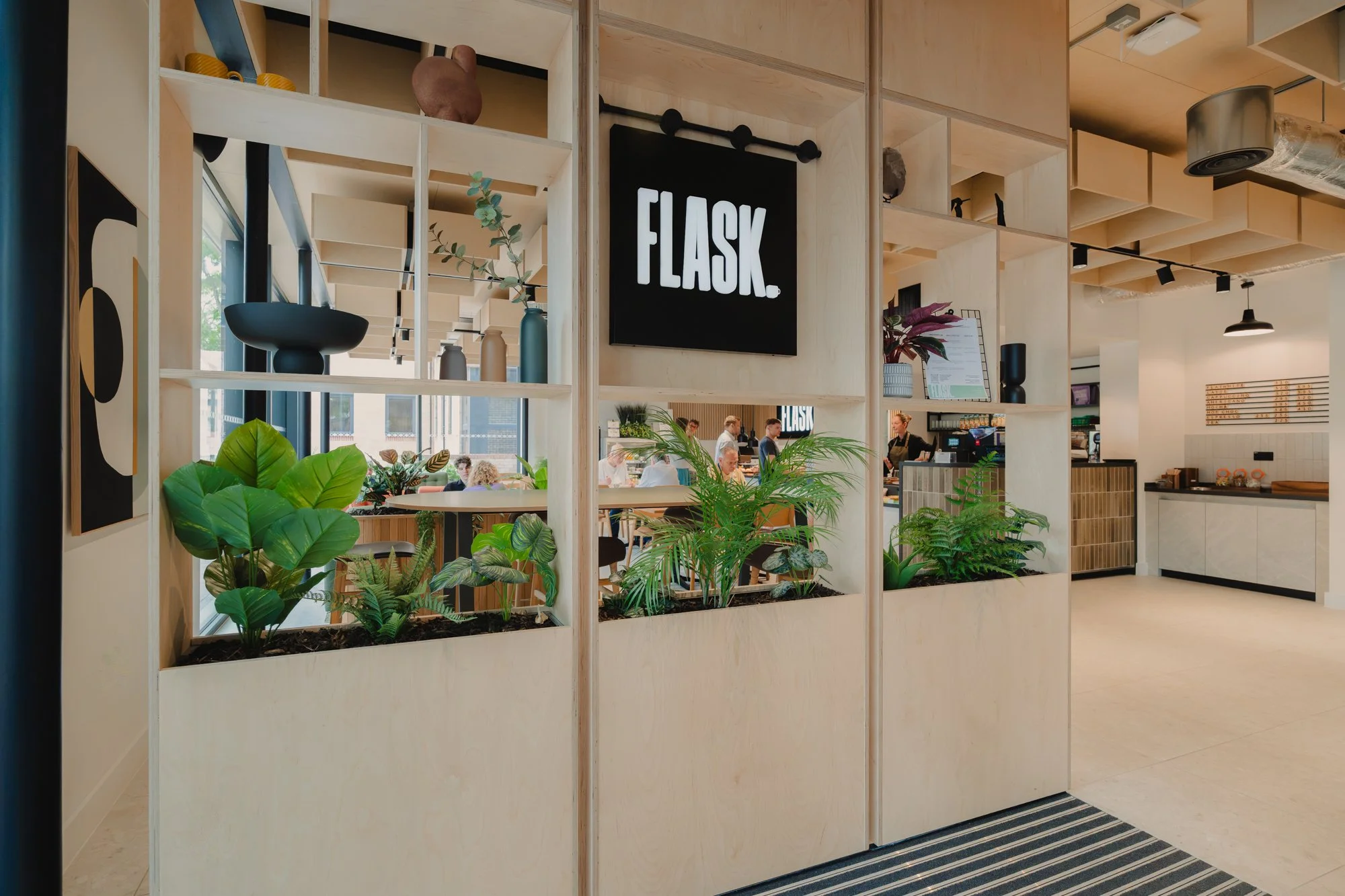 Modern cafe interior with wooden shelving that contains potted plants and decorative items, a black illuminated sign with the word 'FLASK,' and a counter area with menu and staff in the background.