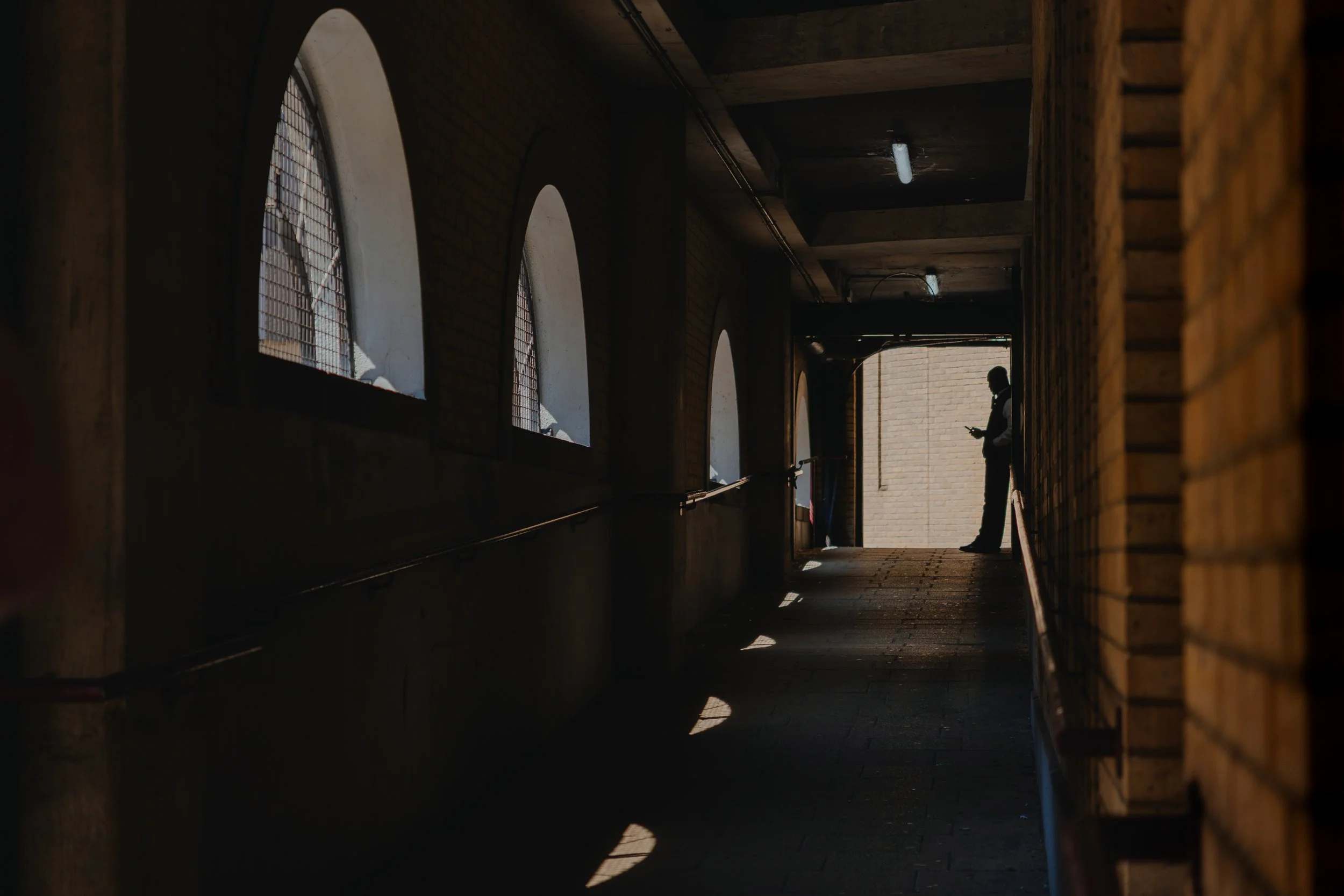 Silhouette of a person standing in a dimly lit corridor, looking at their phone, with sunlight coming from outside at the end of the corridor.