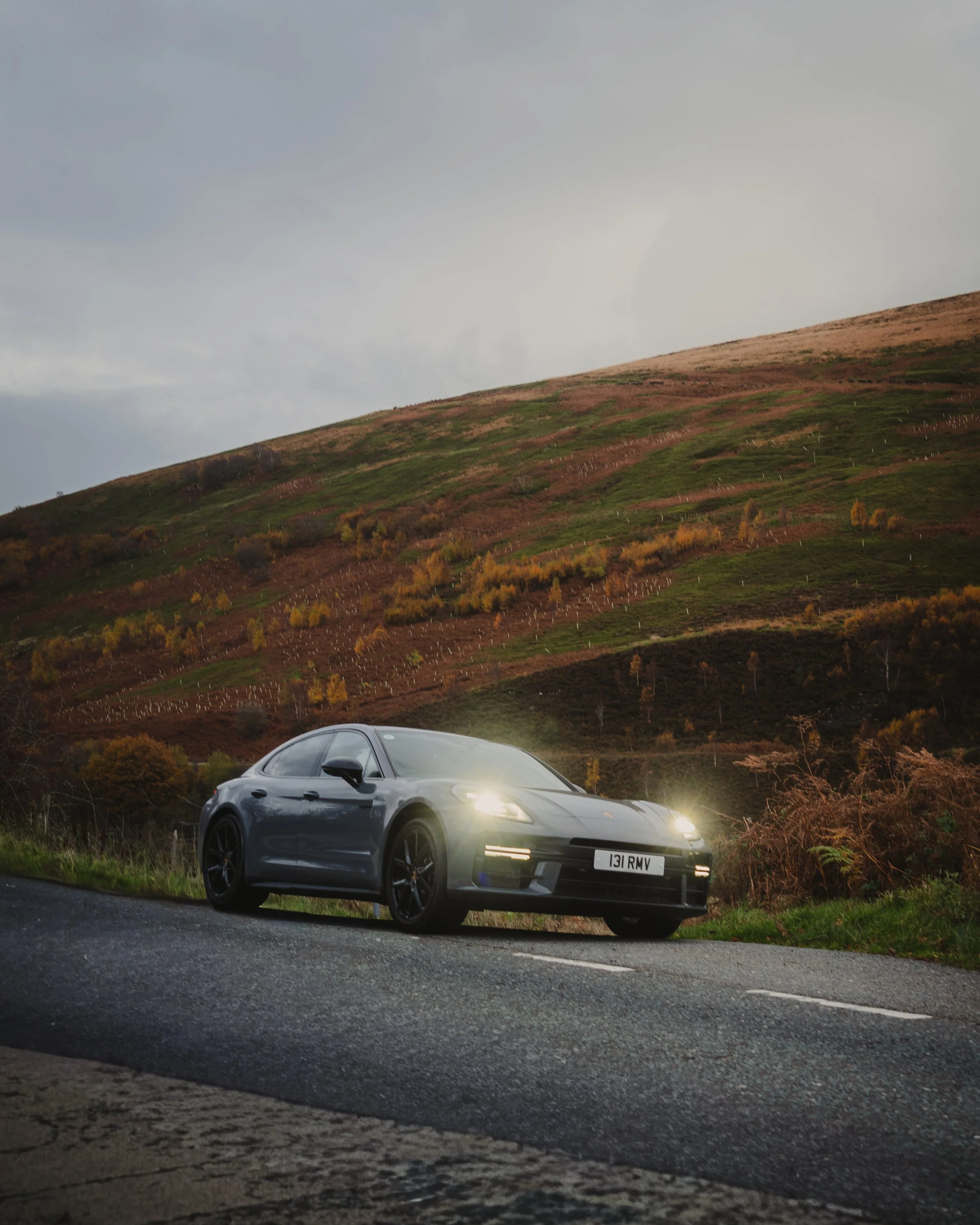A gray sports car with headlights on is parked on the side of a road, with a hillside covered in green and brown landscape in the background.