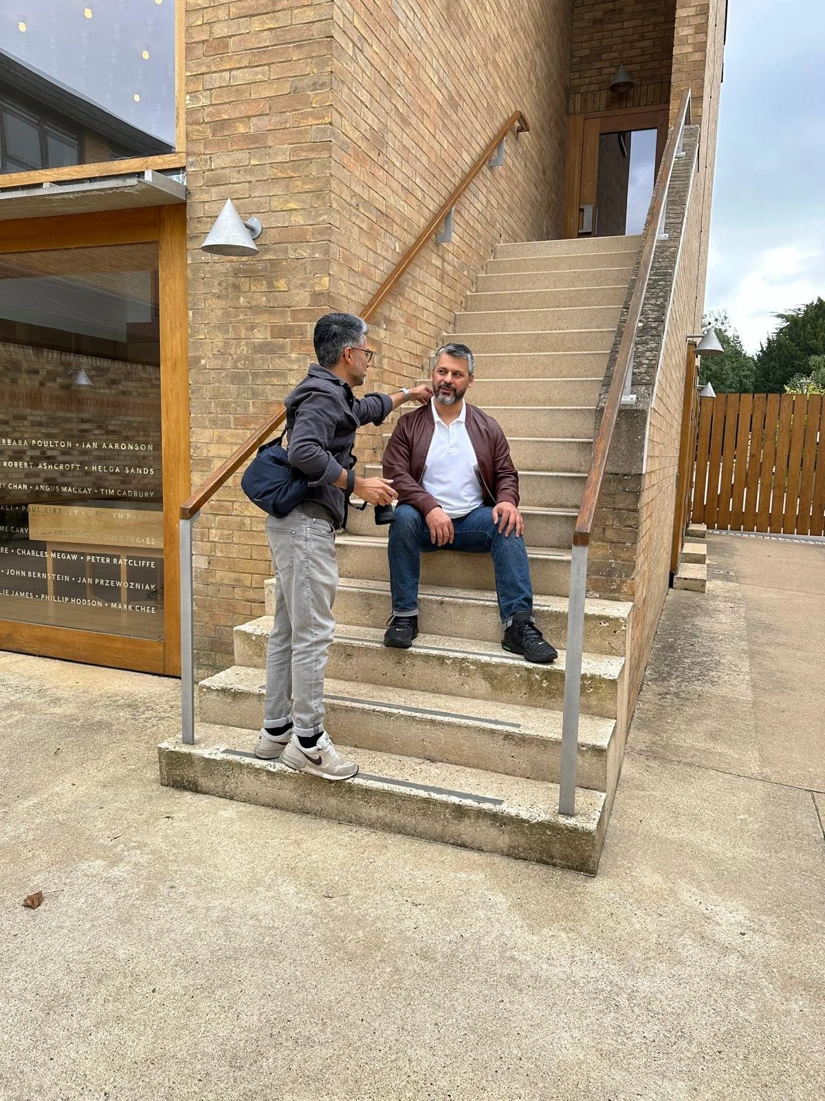 Two men are having a conversation, on concrete stairs outside a brick building, one sitting on the stairs and the other, aa photographer is directing him in the photoshoot, with a wooden handrail and a wooden door in the background.