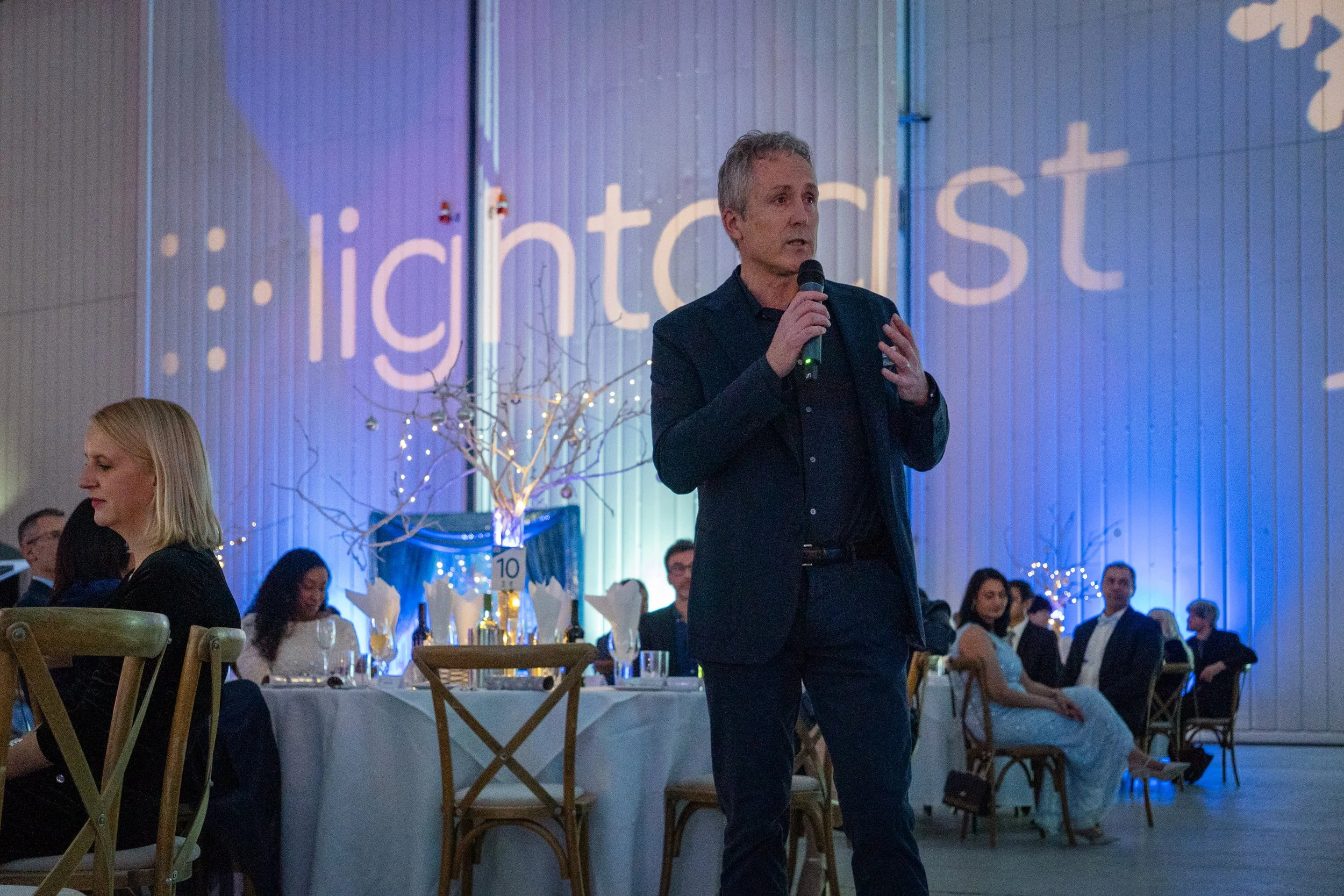 A man in a dark suit holding a microphone, speaking at a formal event with people seated at tables decorated for a celebration.