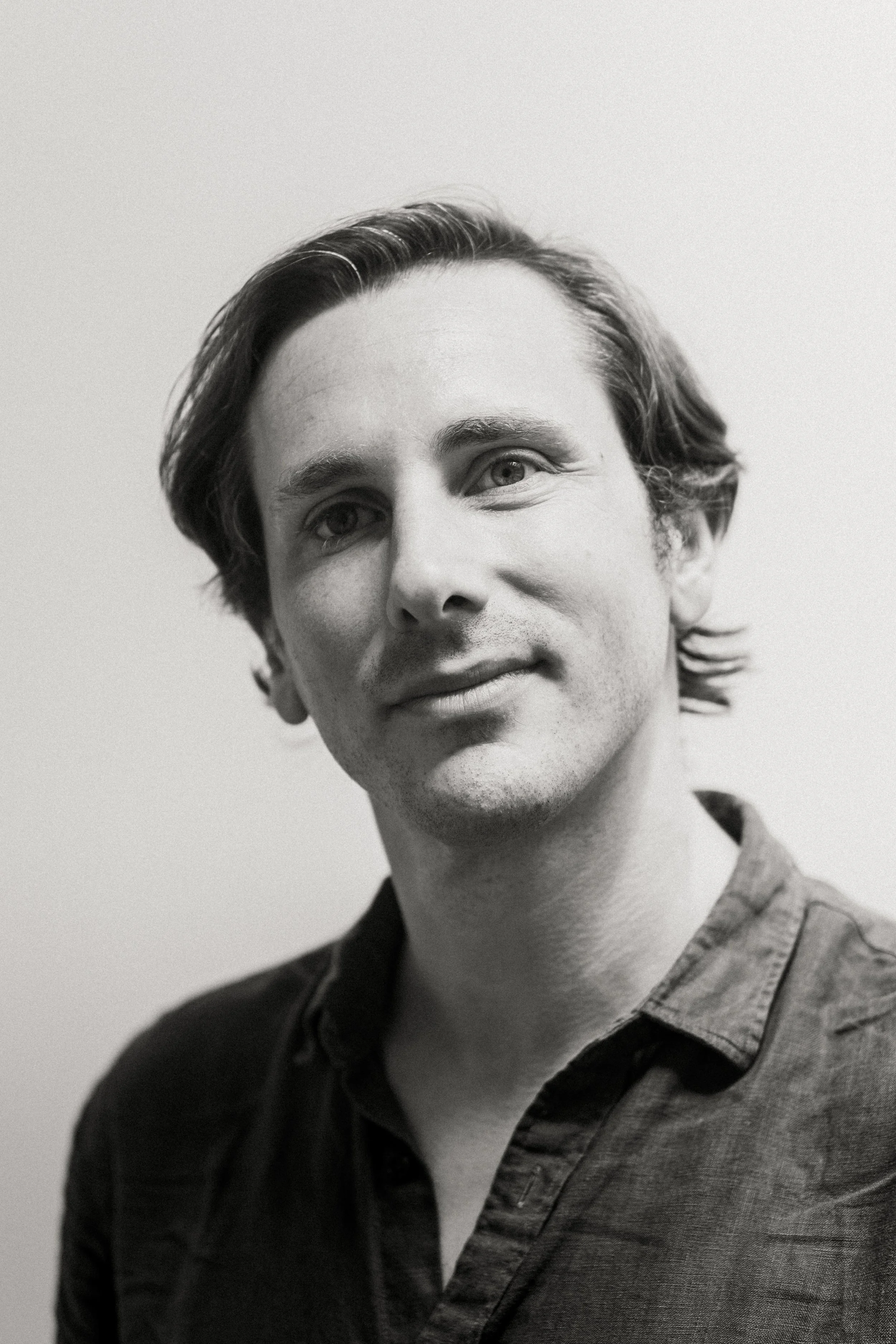 A black and white portrait of a man with medium-length hair, lightly smiling, wearing a collared shirt against a plain background.