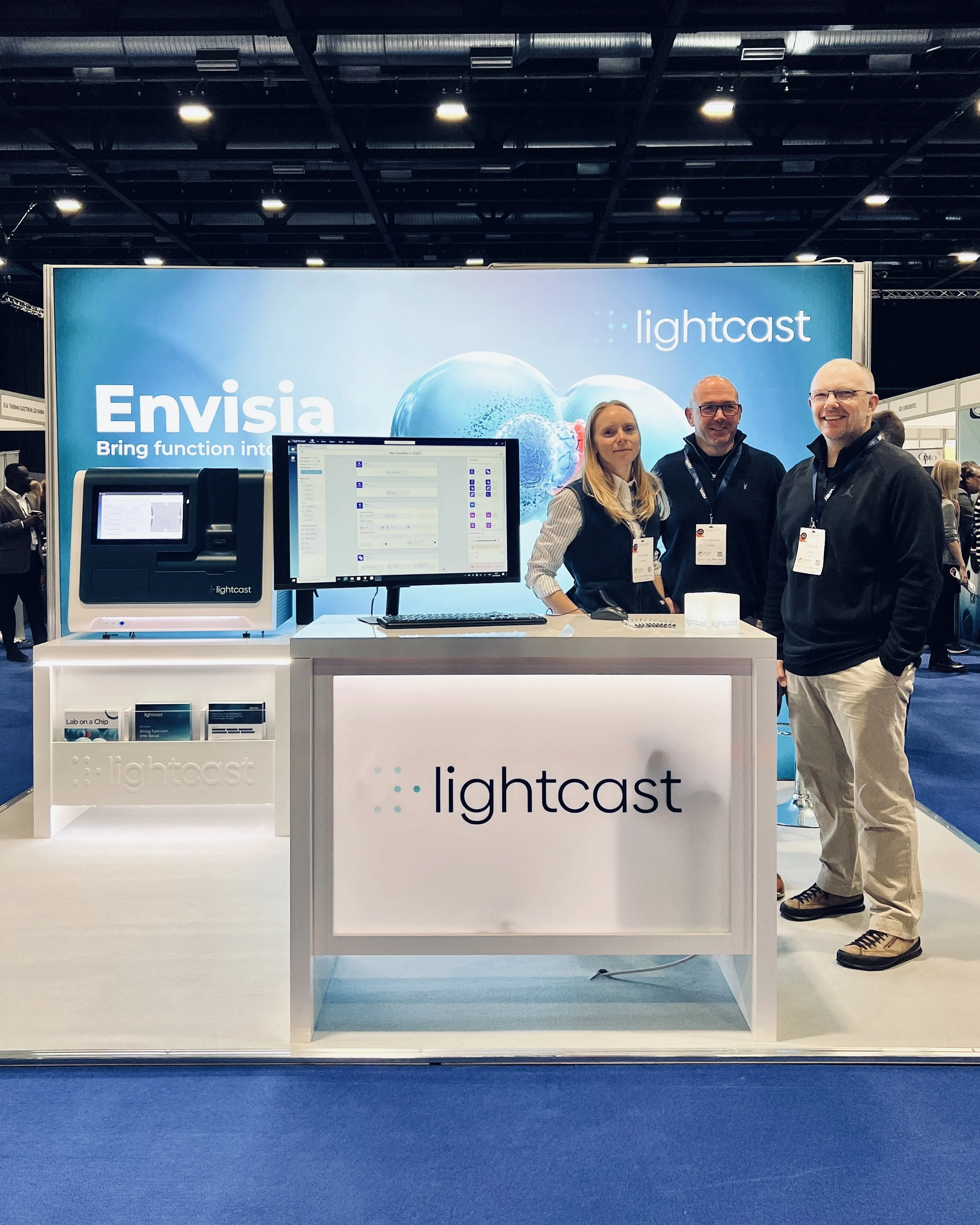 Three people standing behind a white booth with the word 'lightcast' on it, in a convention center with a large blue promotional backdrop behind them displaying the words 'Envisia' and 'Bring function into'. The booth has computer equipment and promo