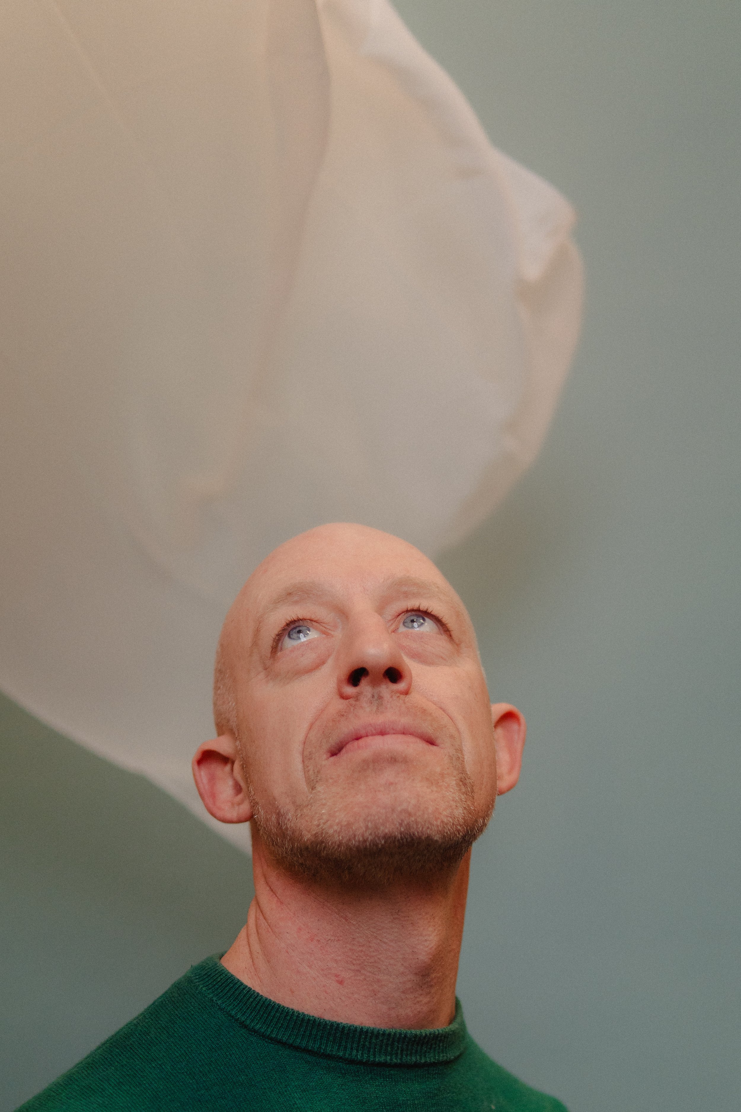 A man with a shaved head and blue eyes looking upward, with a white cloth blowing in the air.