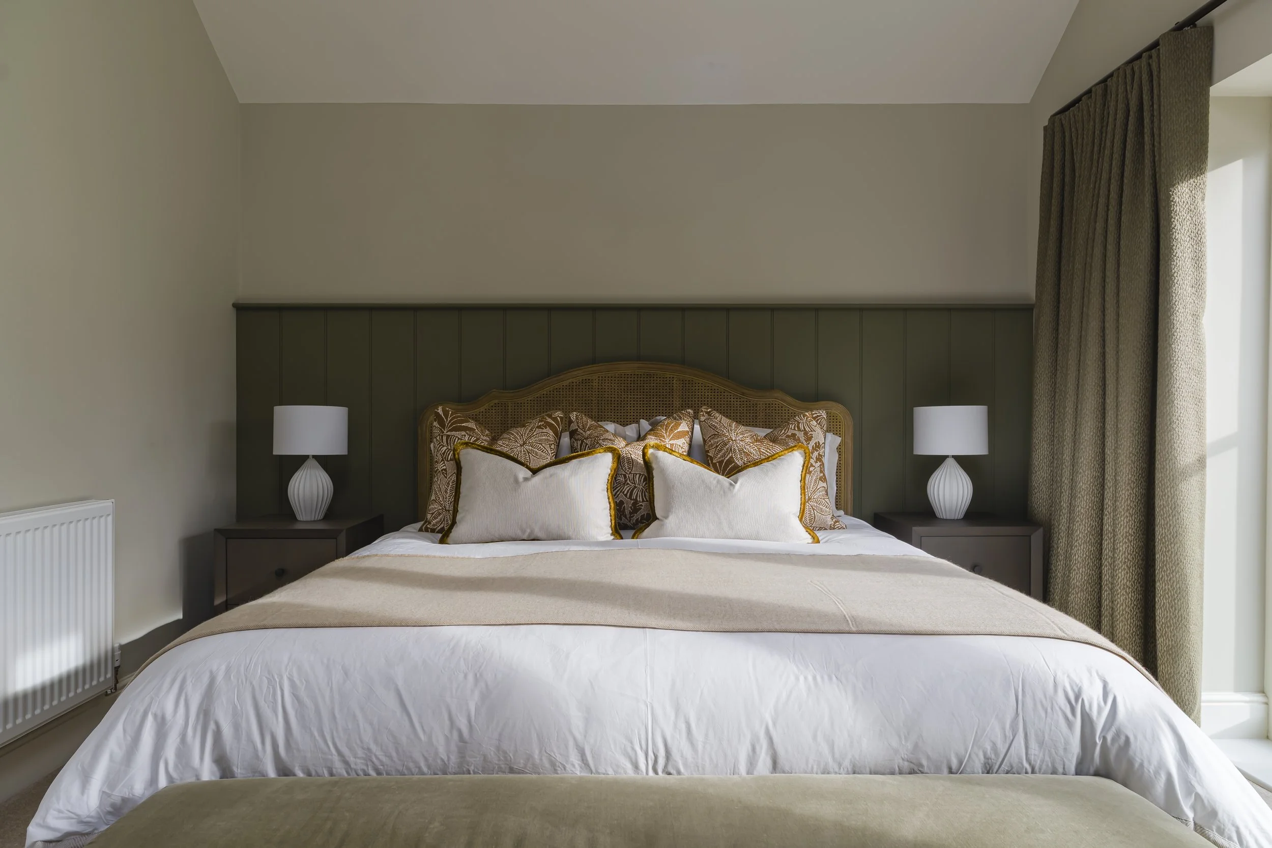 A neatly made bed with decorative pillows in a bedroom with beige and green walls, two nightstands with white lamps, and a window with beige curtains.