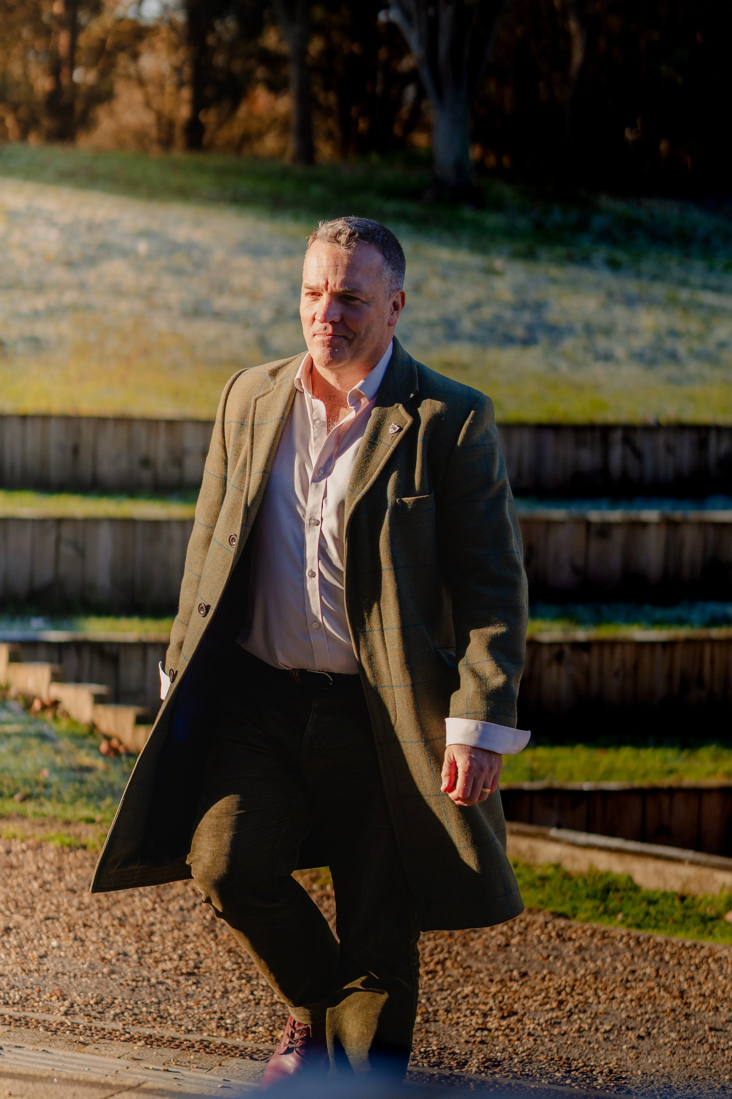 A man wearing a brown plaid coat, white shirt, and dark trousers walking outdoors on a sunny day with trees in the background.